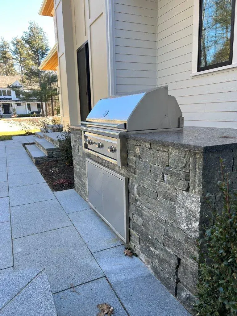 Outdoor stainless steel built-in grill beside a stone wall along a paved patio walkway