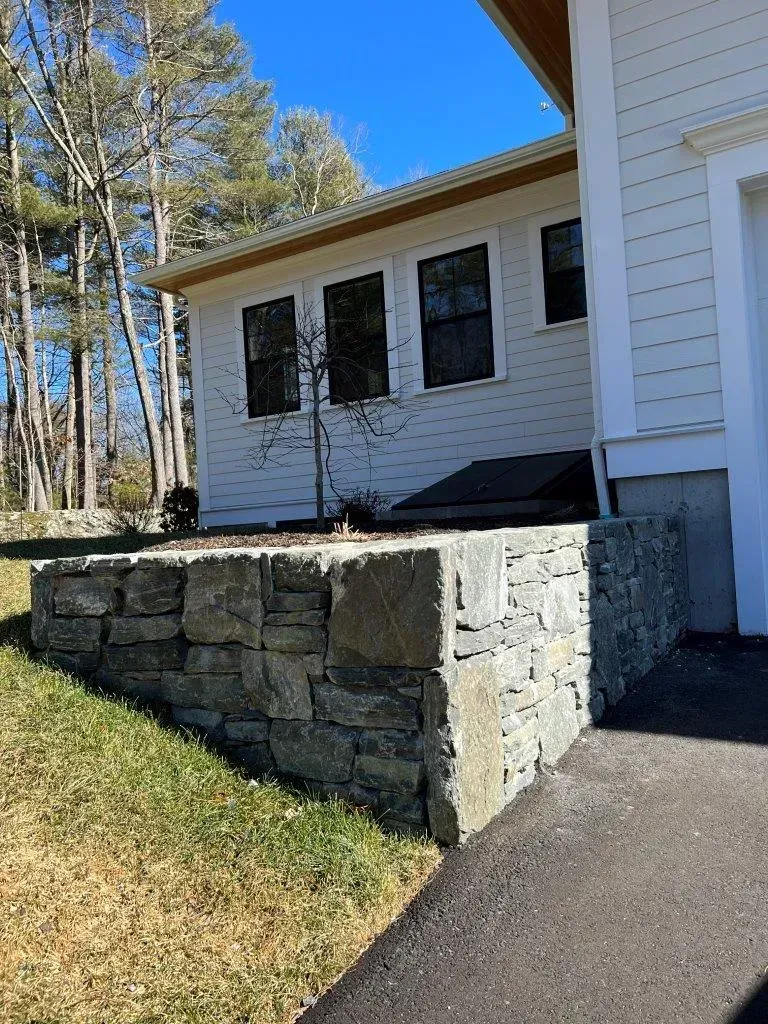 Stone retaining wall beside a white house with black shutters and a paved walkway