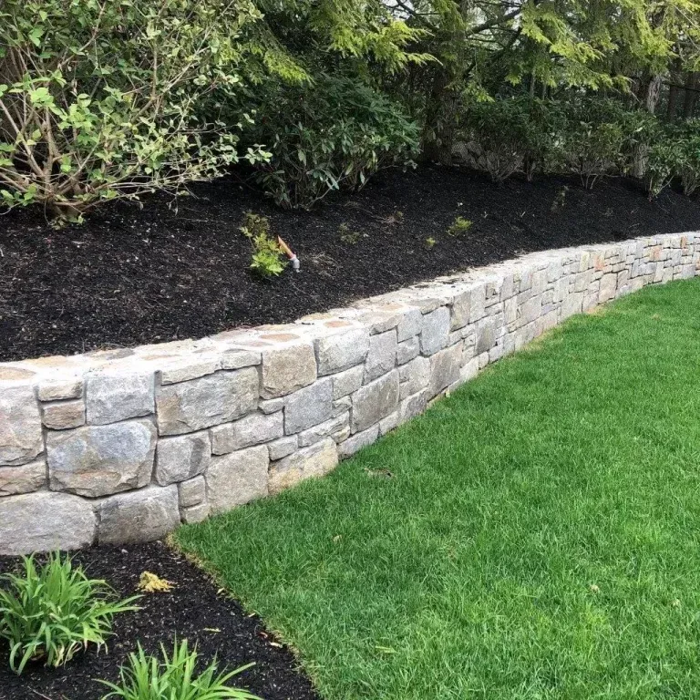 Curved stone retaining wall beside a green lawn and mulched garden bed with shrubs