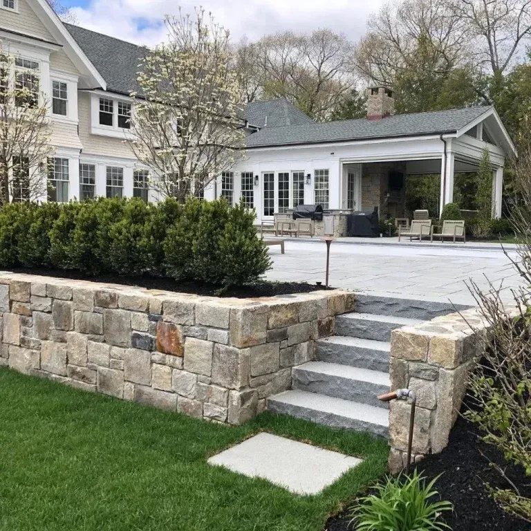 Stone retaining wall with steps leads to a white house patio and covered porch in a landscaped yard