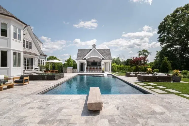 Modern backyard pool with stone patio, lounge chairs, and a large white house under a bright sky