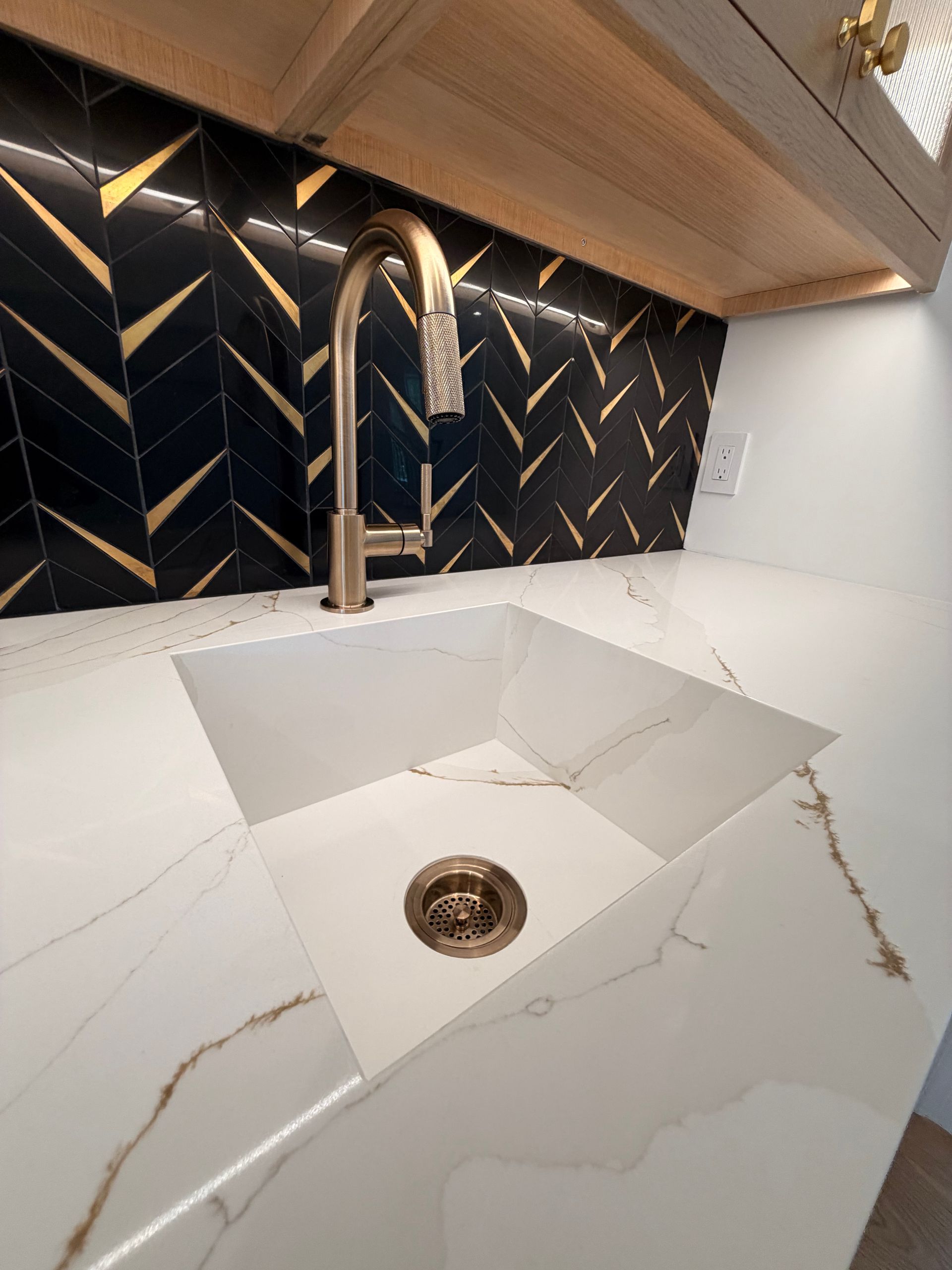 Modern kitchen sink with white countertop, brass faucet, and black geometric tile backsplash