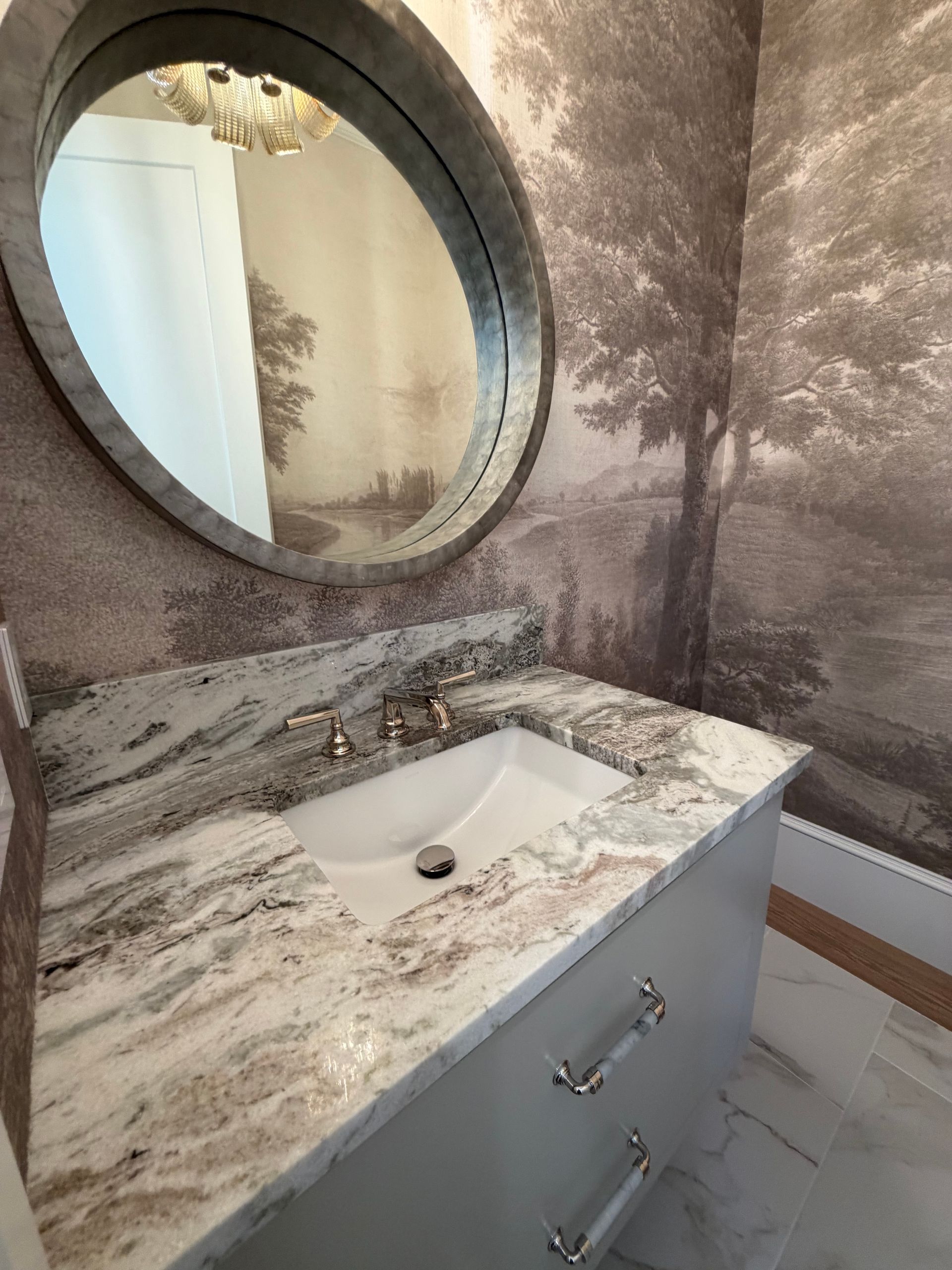 Small bathroom vanity with marbled countertop, oval mirror, and chrome faucet against patterned wallpaper.