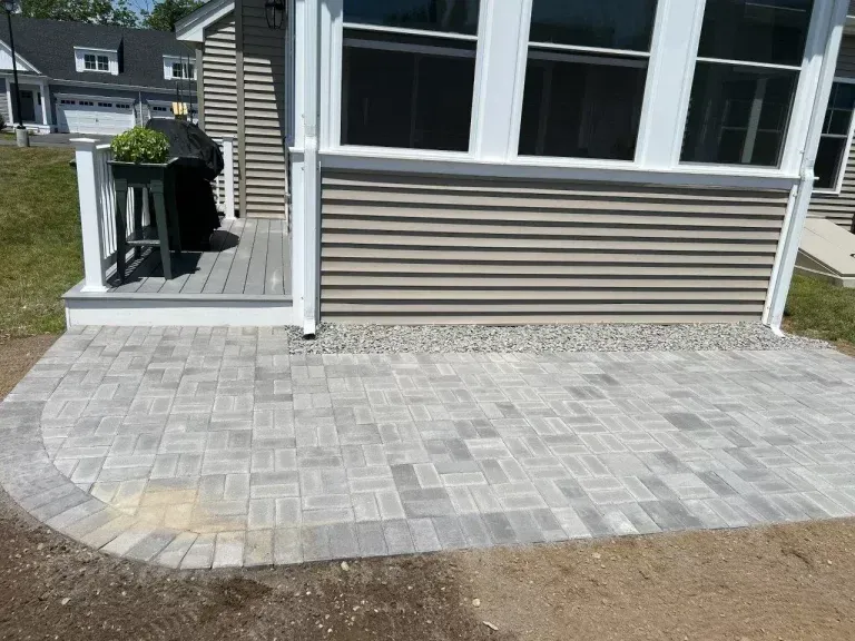 Paver patio outside a house with large windows and a side walkway to a deck.