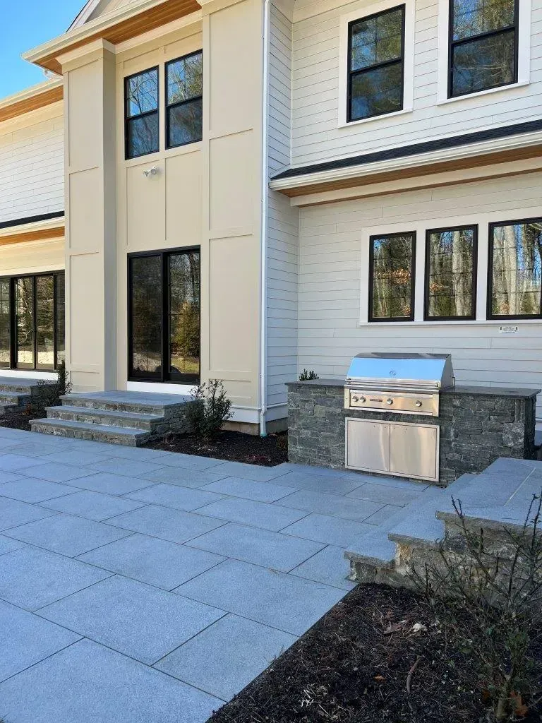 Modern house exterior with stone patio, white siding, black windows, and stainless steel grill on the side