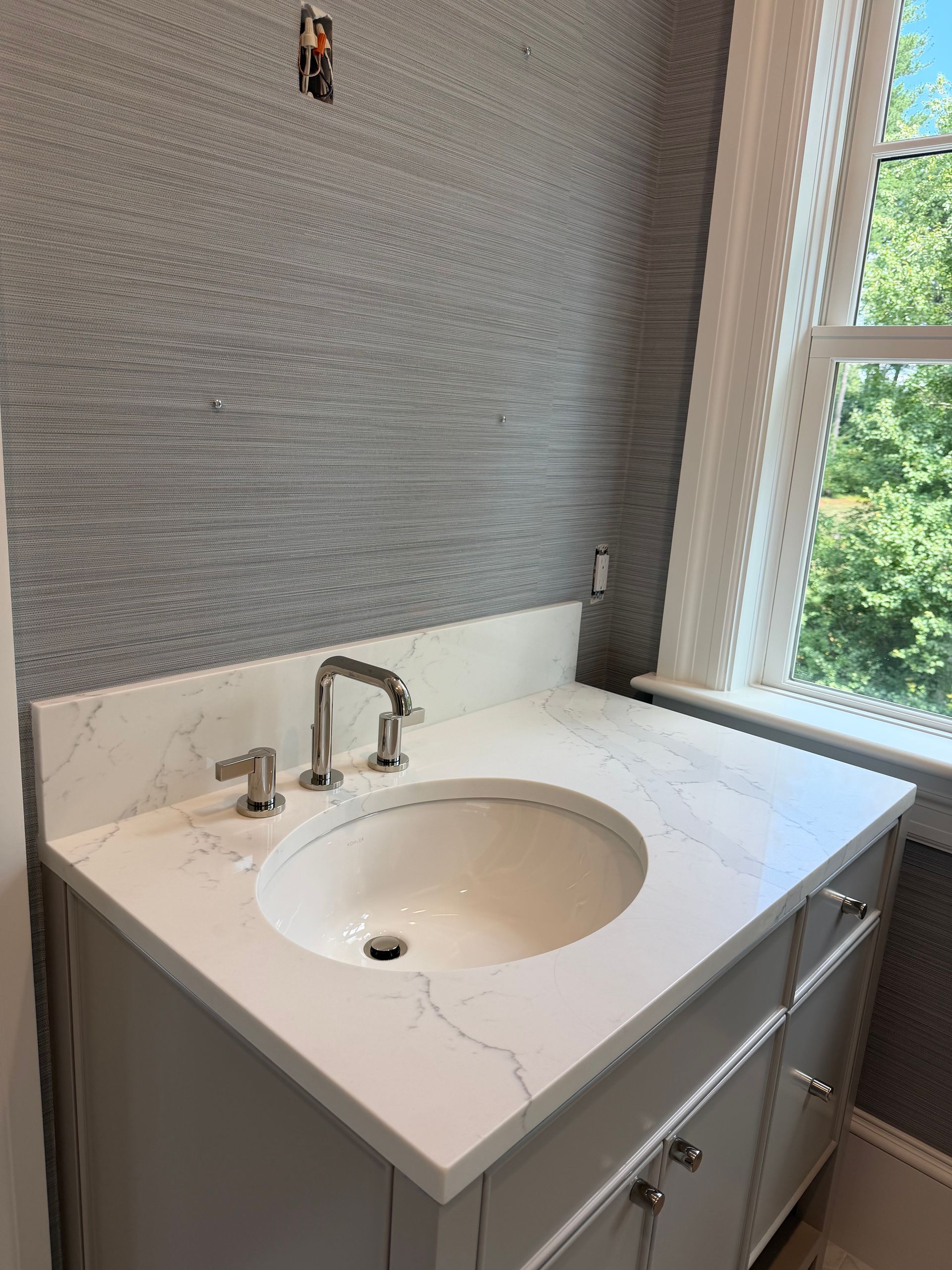 Bathroom vanity with white sink, chrome faucet, gray textured wall, and window beside the counter