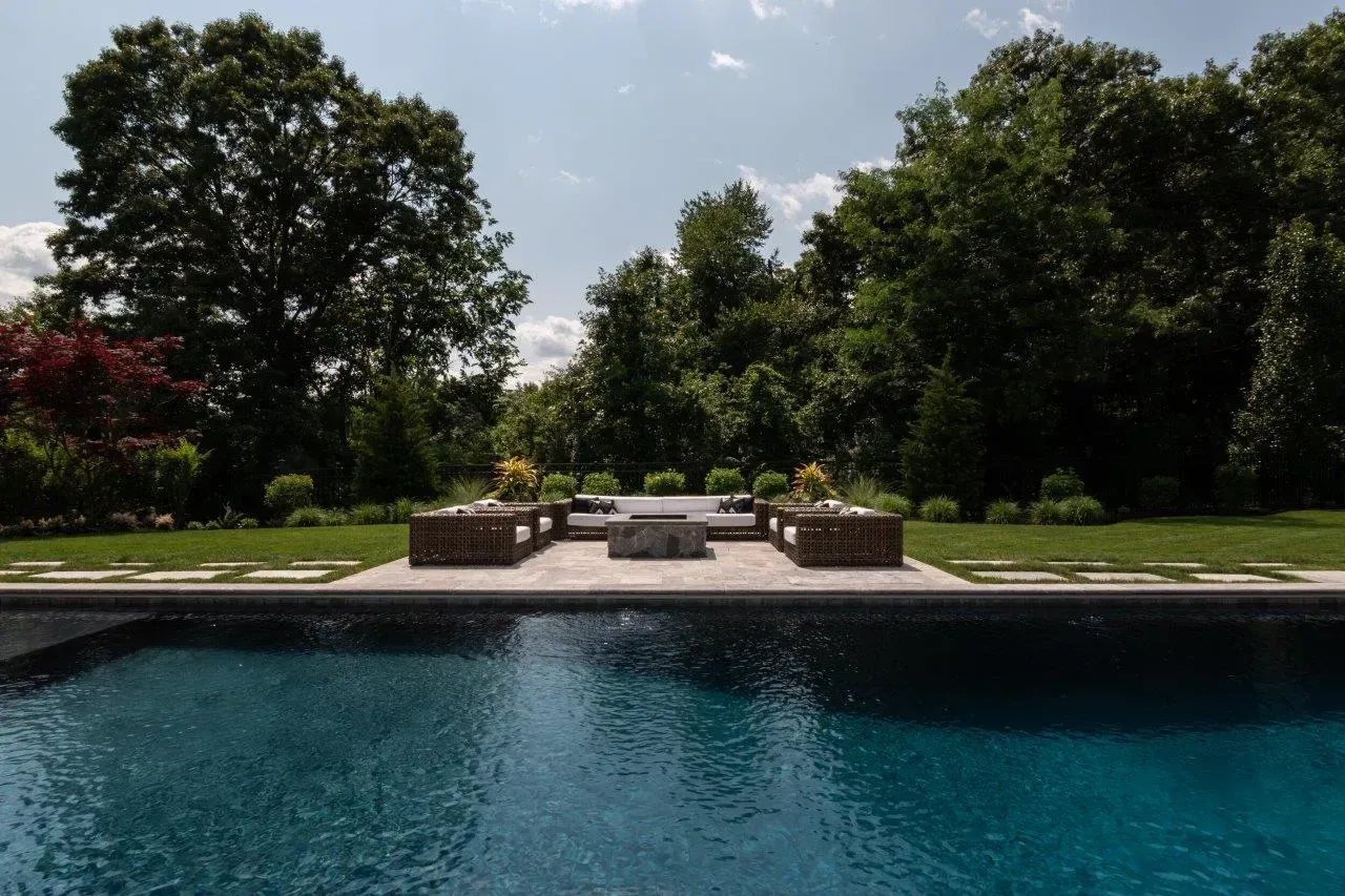 Poolside lounge chairs beside a turquoise pool, with lush green trees and a bright sky beyond.