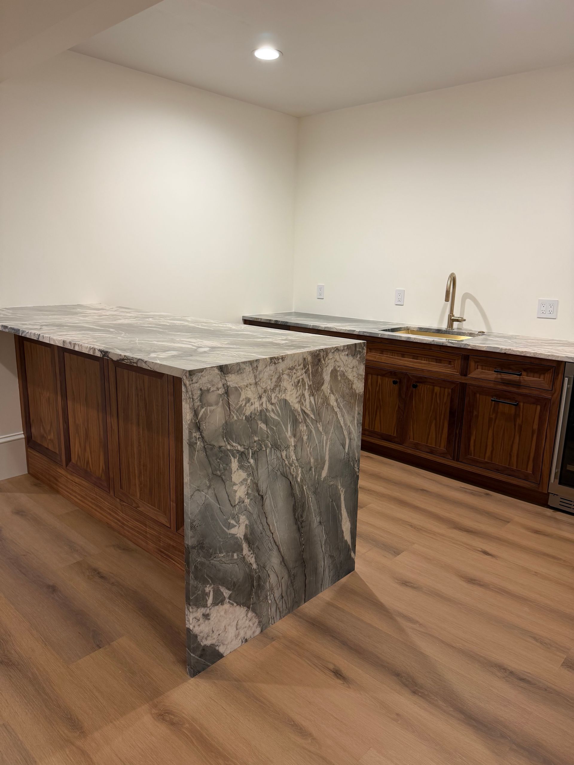 Empty kitchen with a large marble island, wood cabinets, and stainless sink under bright ceiling lights
