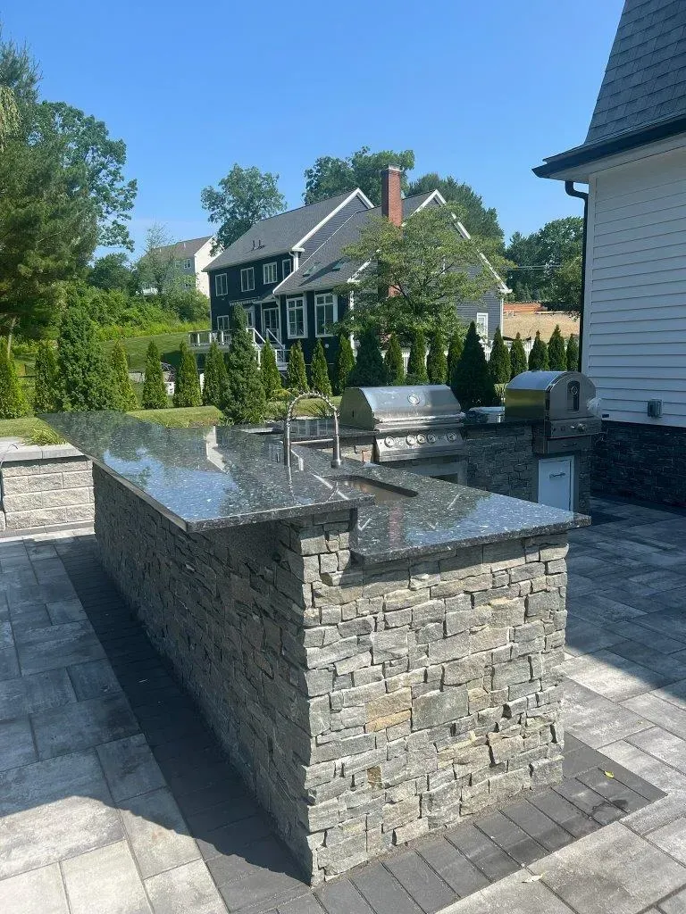 Stone outdoor kitchen island in a landscaped backyard beside a house, with trees and hedges in the background