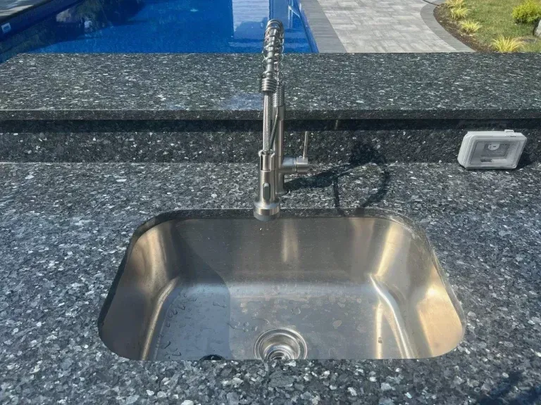 Stainless steel sink with tall faucet beside a blue pool and stone countertop outdoors
