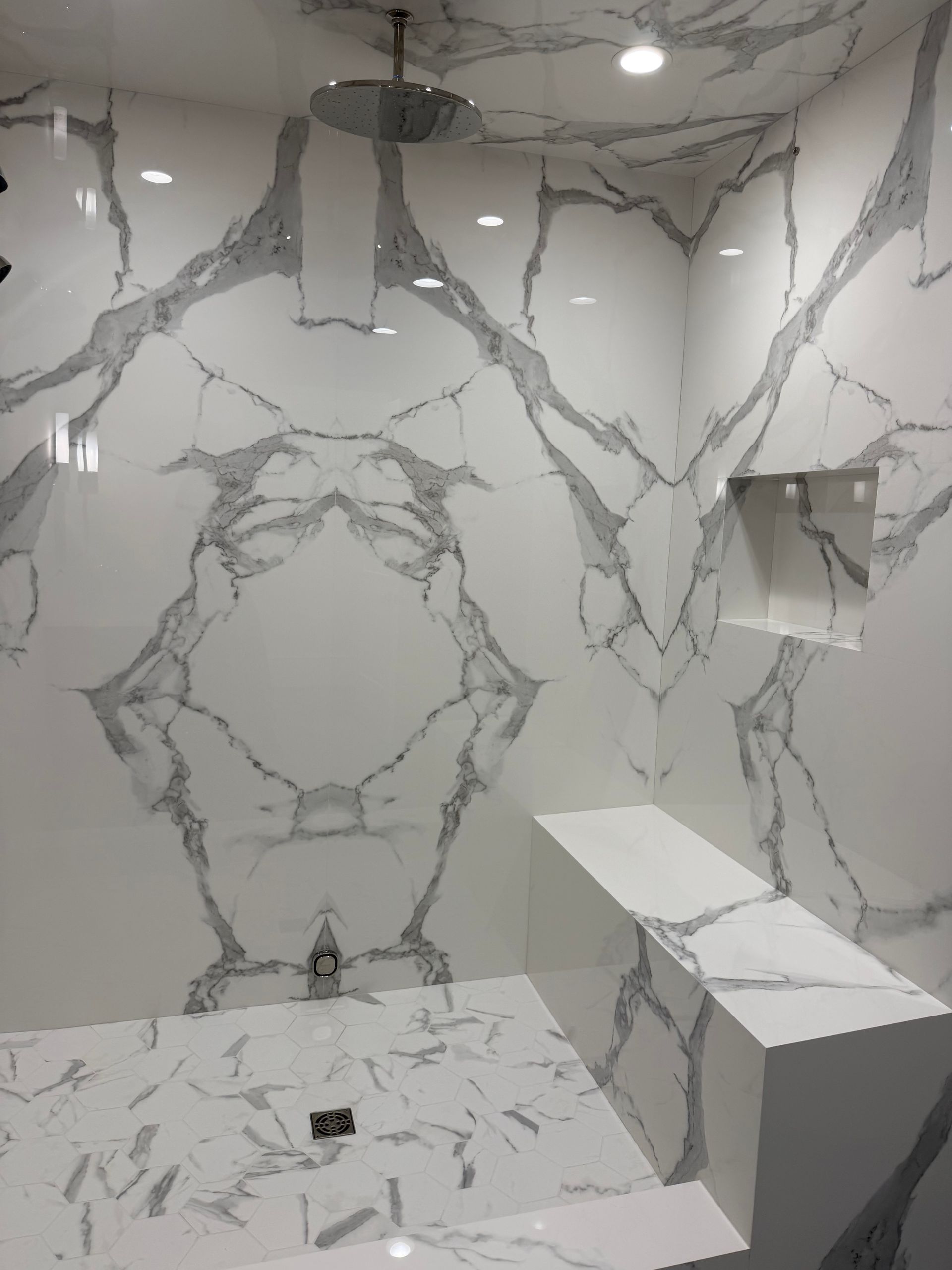Marble-tiled shower with a ceiling showerhead, bench, recessed niche, and floor drain