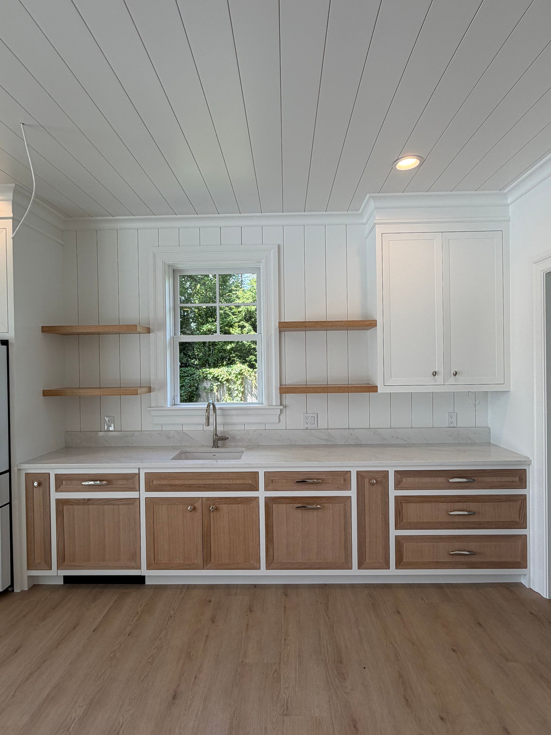 Bright white kitchen with wood cabinetry, open shelves, and a central window over the sink.