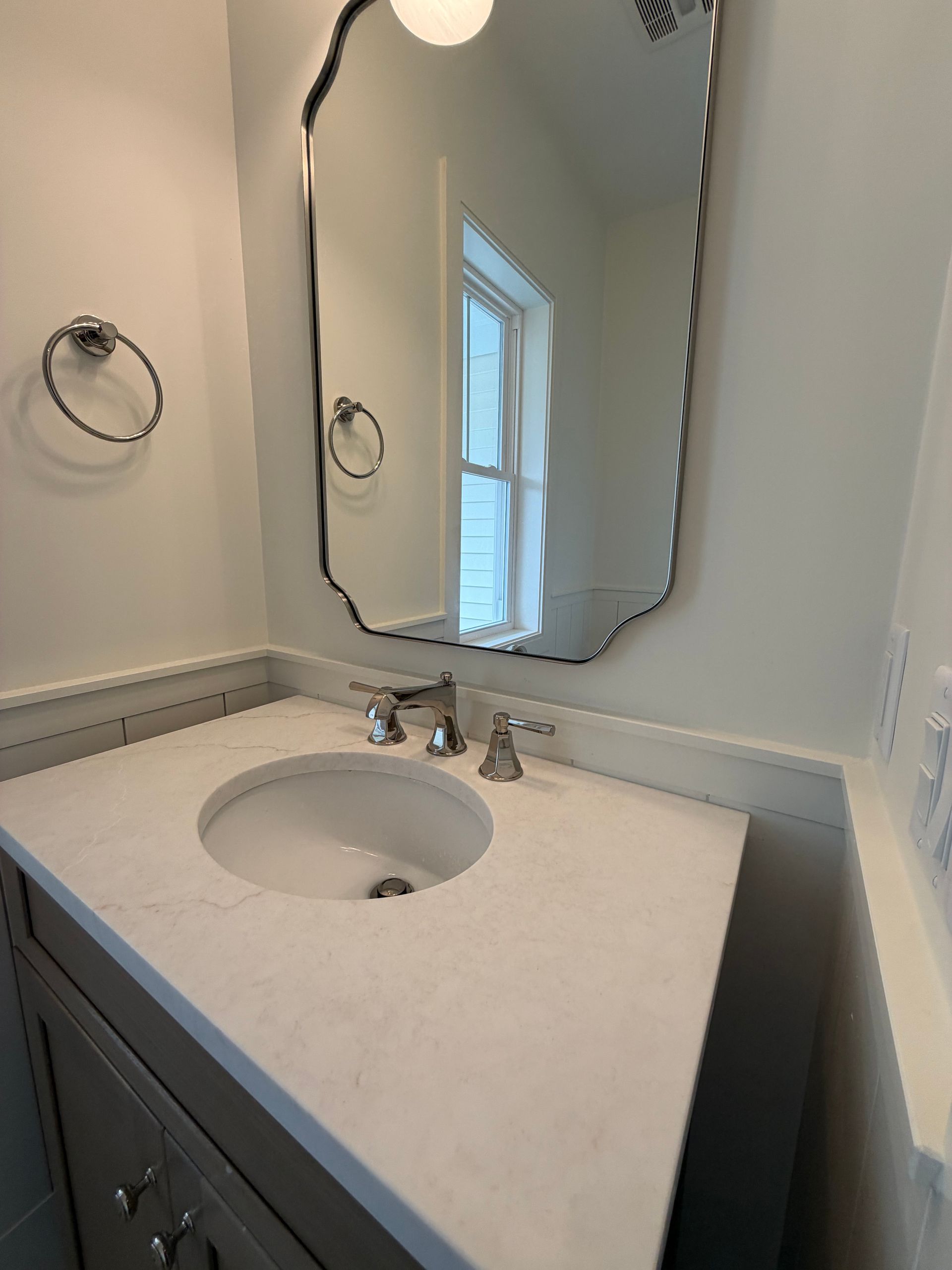 Small bathroom vanity with white countertop, oval sink, chrome faucet, and wall mirror beside a window.