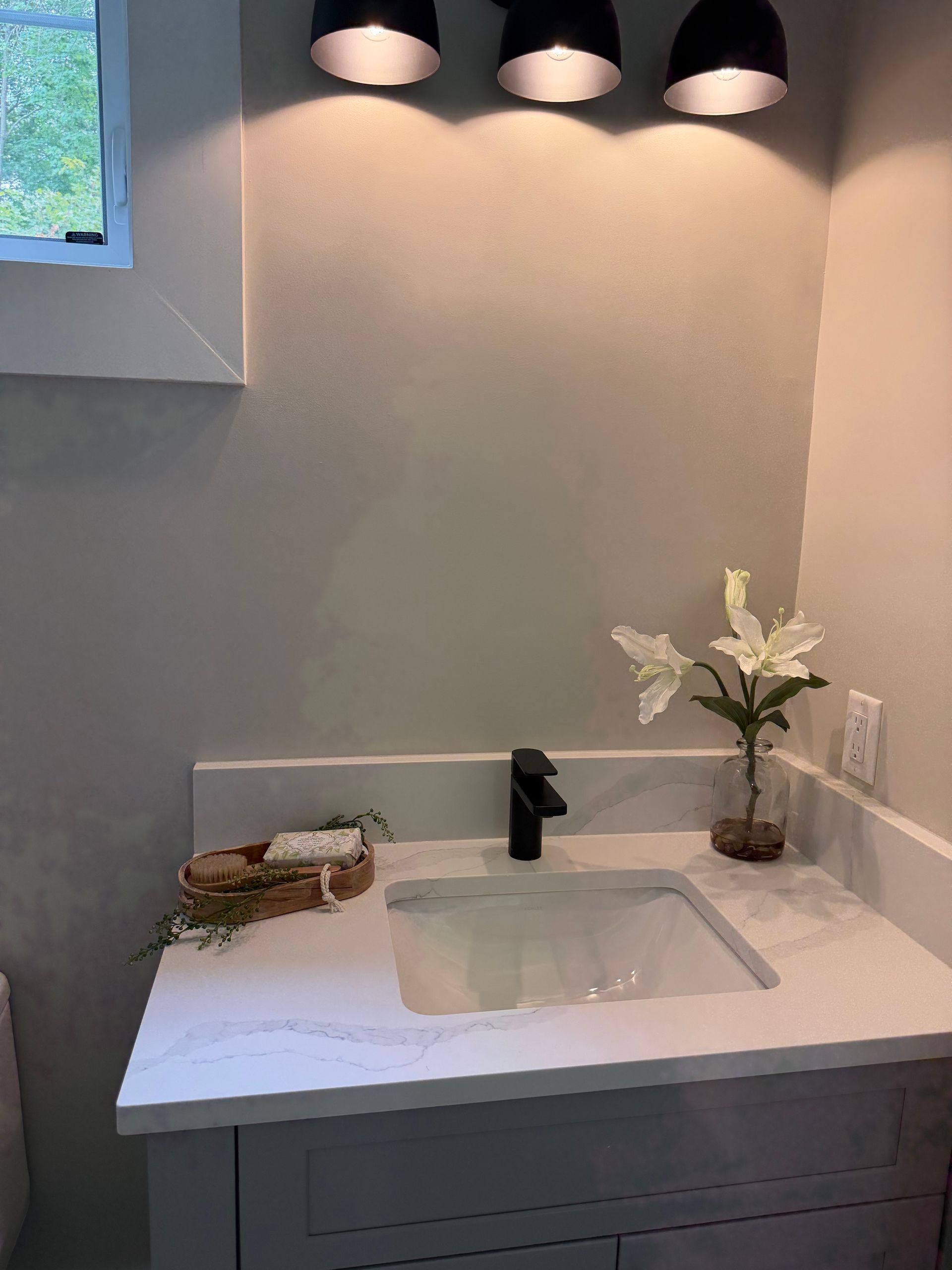 Minimal bathroom vanity with white sink, black faucet, candle, and vase of flowers under three wall lights.
