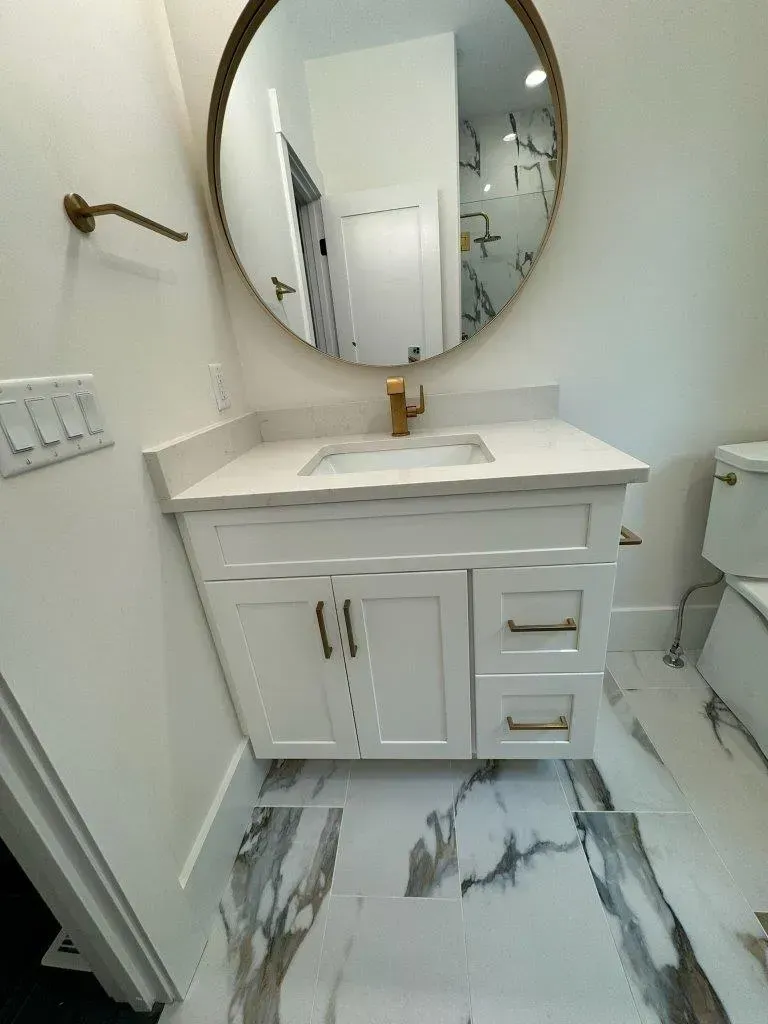 Small bathroom vanity with round mirror, gold faucet, white cabinetry, and marble-pattern tile floor.