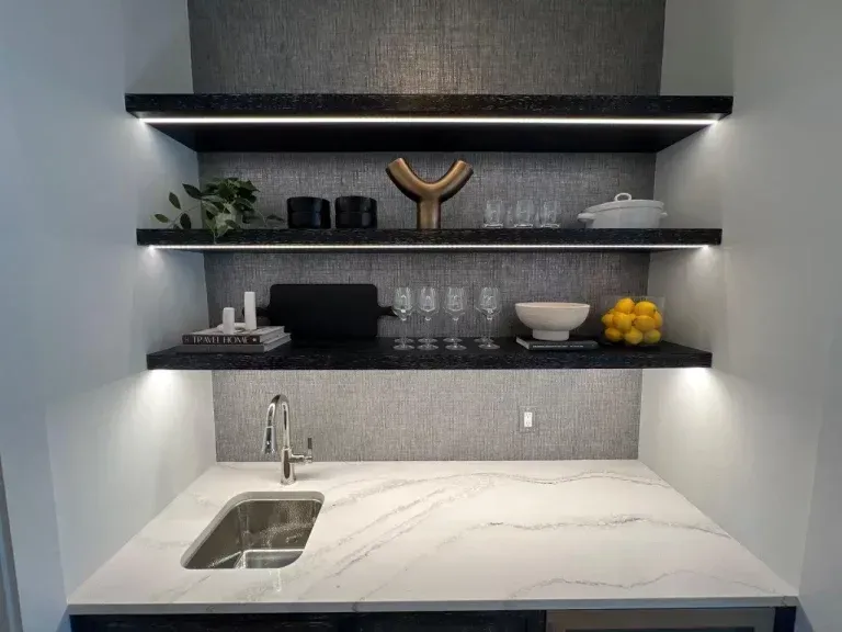 Modern kitchen with marble counters, floating shelves, sink, black accents, and fruit bowl on the right.