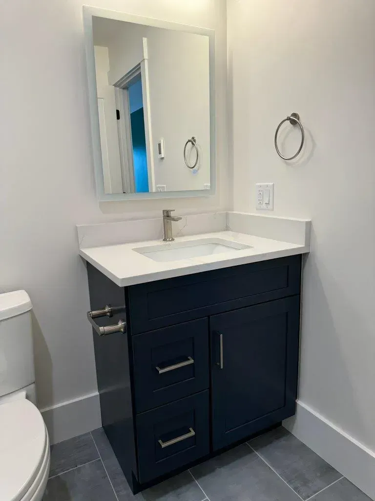 Small bathroom vanity with navy cabinet, white countertop, mirror, towel rings, and toilet edge visible.