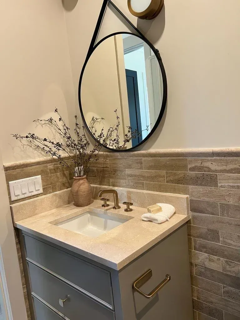 Small bathroom vanity with round mirror, marble countertop, and brass faucet in a tiled alcove.