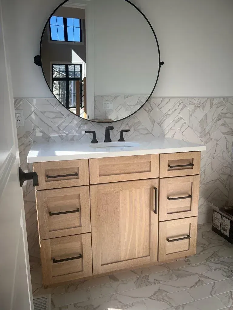 Bathroom vanity with round mirror, double sink, and light wood cabinets in a tiled room