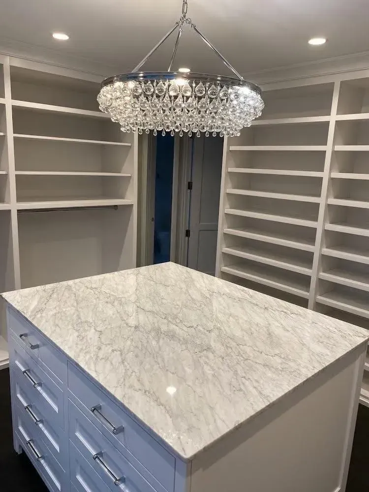 Walk-in closet with white built-in shelves and blue island topped by marble, lit by a crystal chandelier