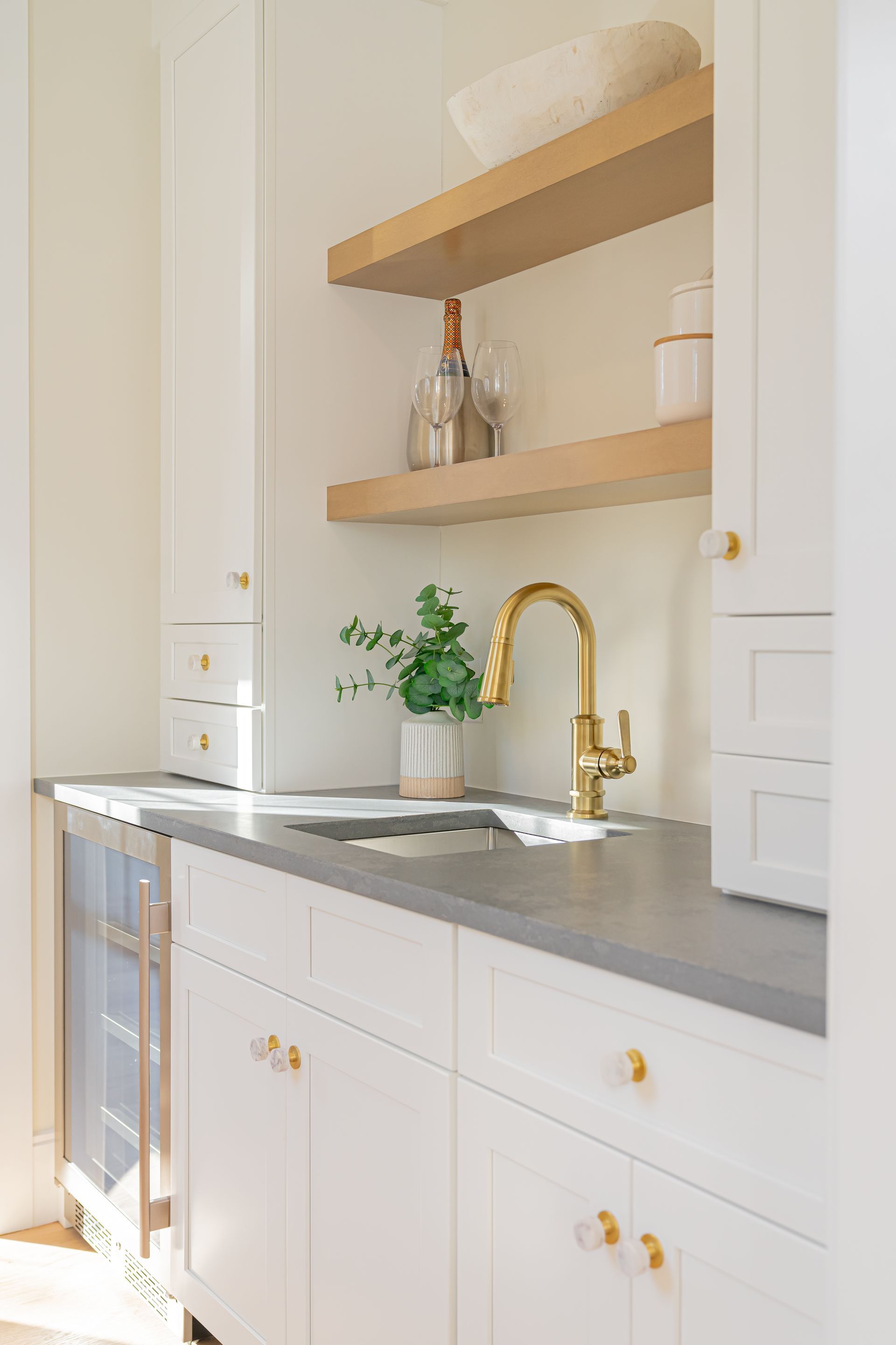 Bright kitchen nook with white cabinets, gray countertop, brass faucet, and wooden shelves with decor and a plant
