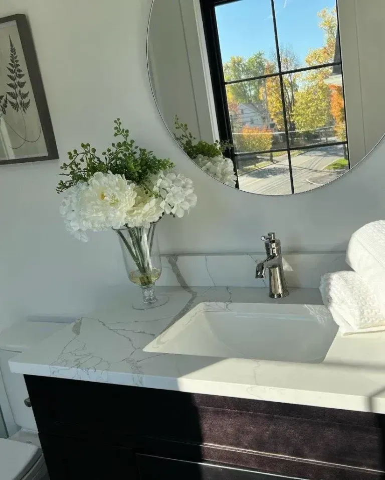 Modern bathroom vanity with round mirror, white flowers, and sunlight through a window