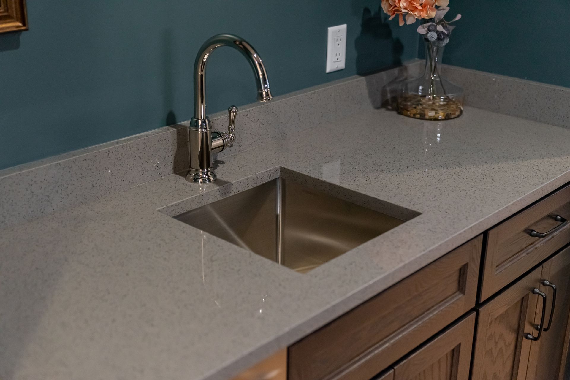 Kitchen countertop with stainless steel sink, chrome faucet, and speckled gray surface against teal walls.