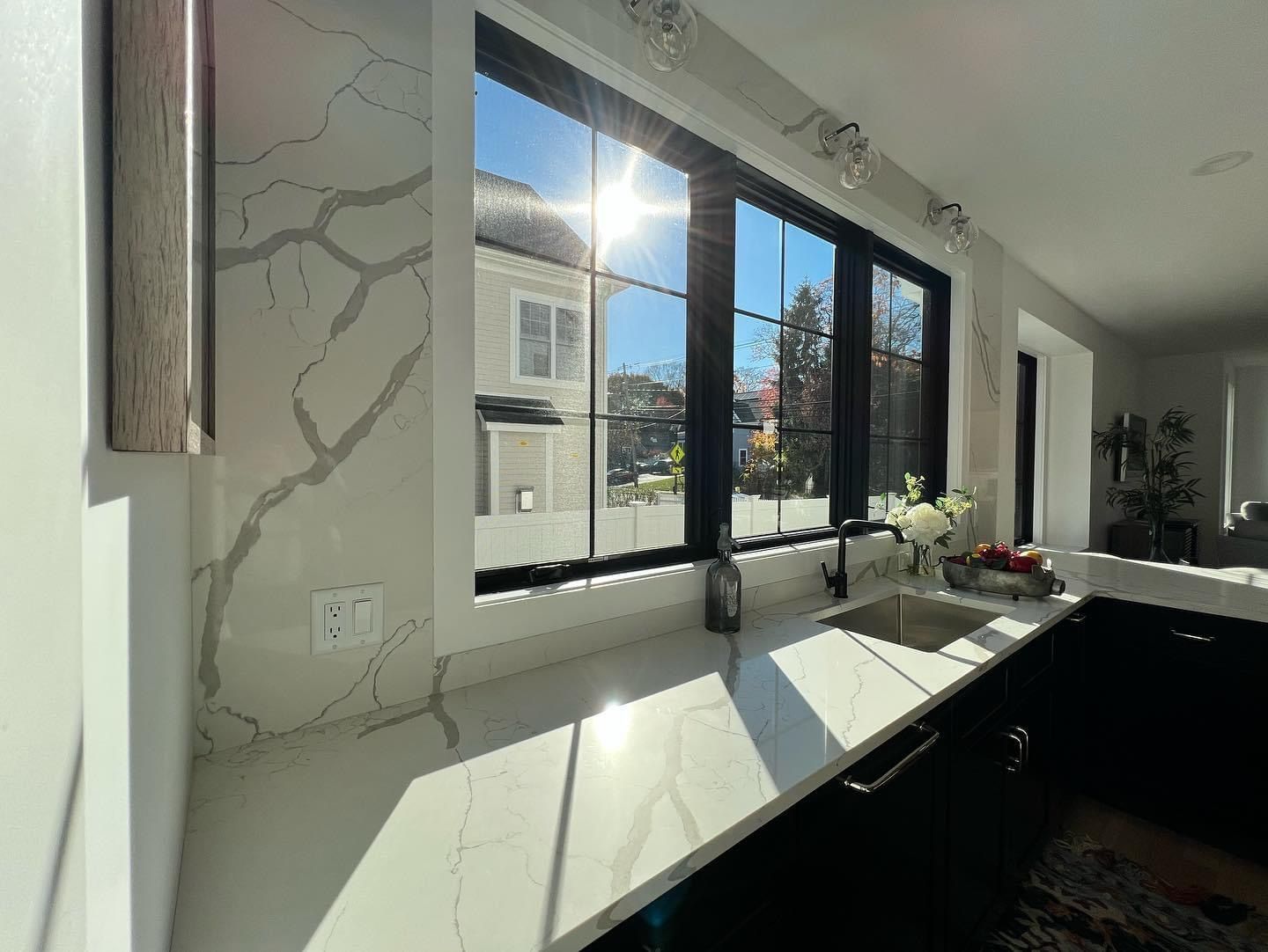 Bright modern kitchen with large black-framed windows, white countertops, and sunlight streaming in.