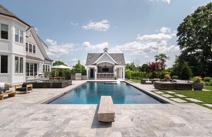 Luxury backyard with a large rectangular pool, stone patio, and elegant white house under a bright blue sky