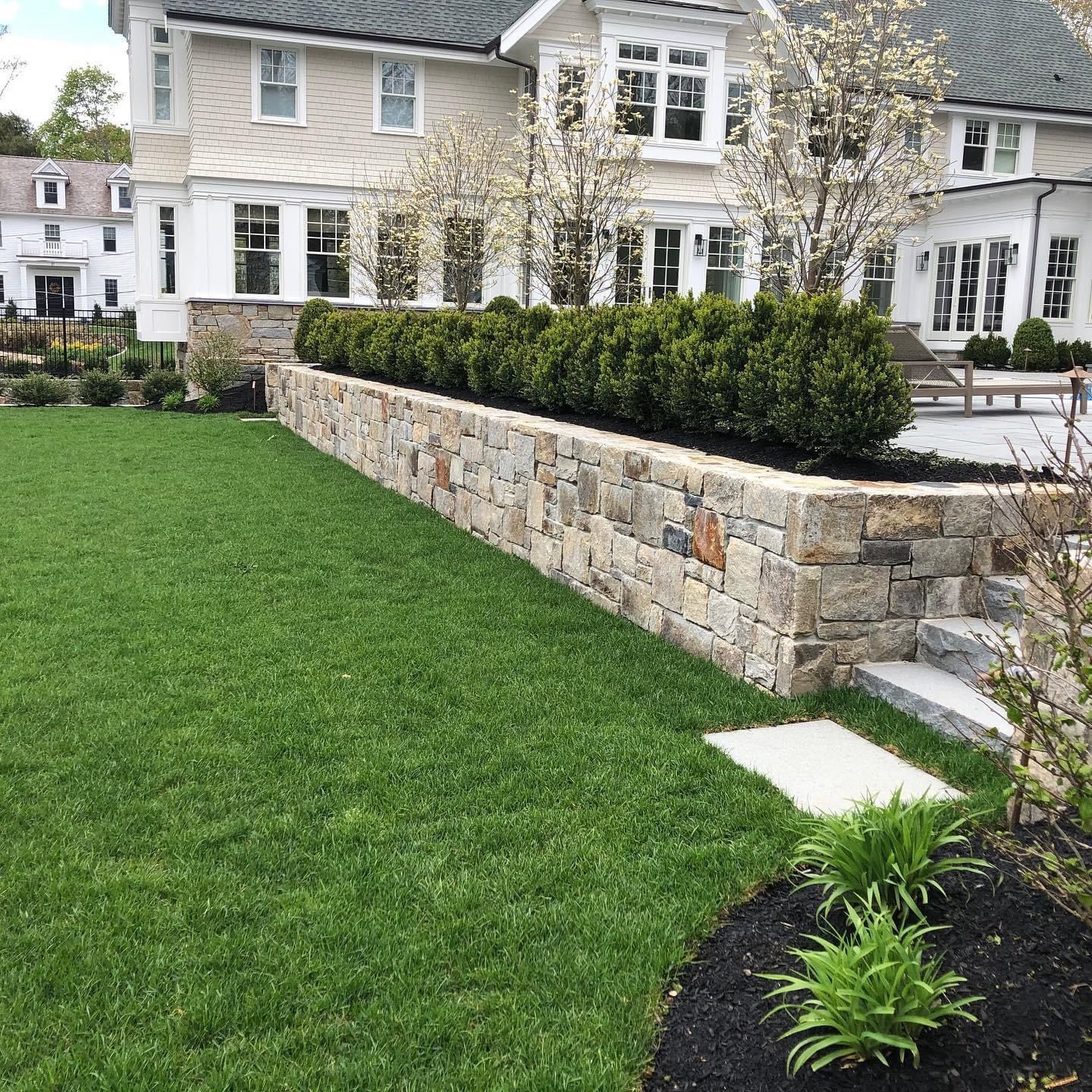 Stone wall and manicured lawn in front of a large stone house with white-trimmed windows and flowering trees