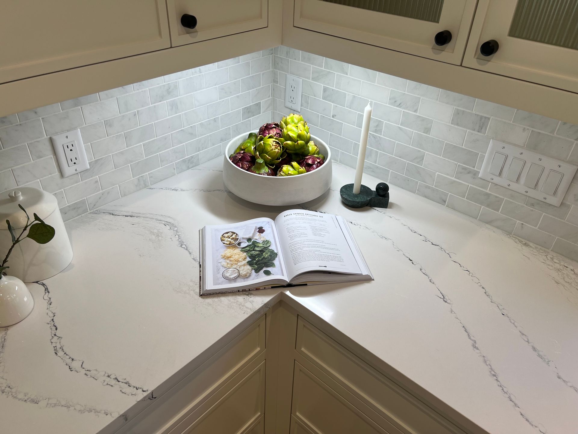 Open cookbook on a white kitchen countertop beside a bowl of fruit and a sink faucet