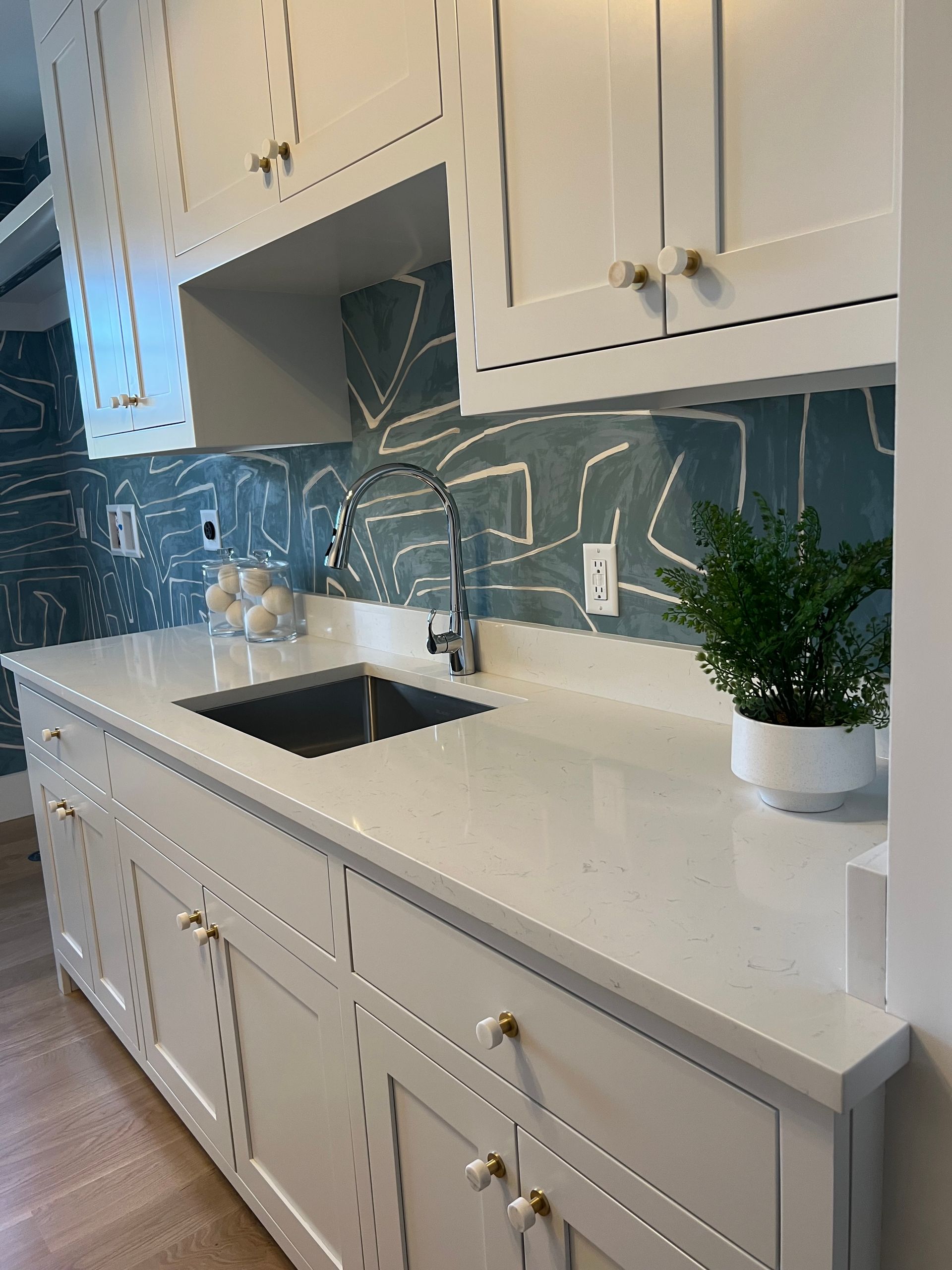 Modern white kitchen with cabinets, stone countertop, black sink, blue tile backsplash, and a potted plant.