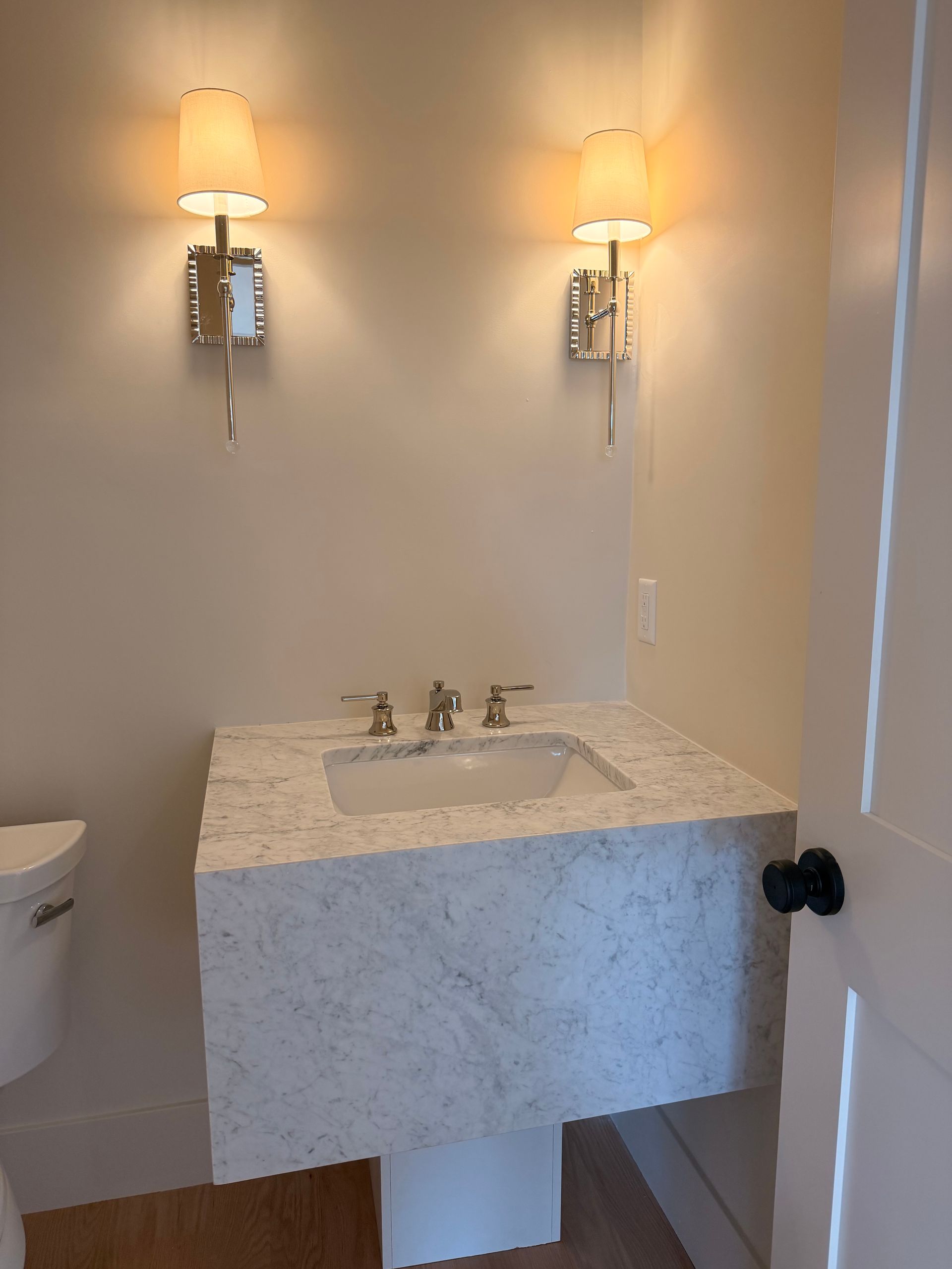 Small bathroom vanity with marble sink, wall sconces, and a toilet visible on the left.