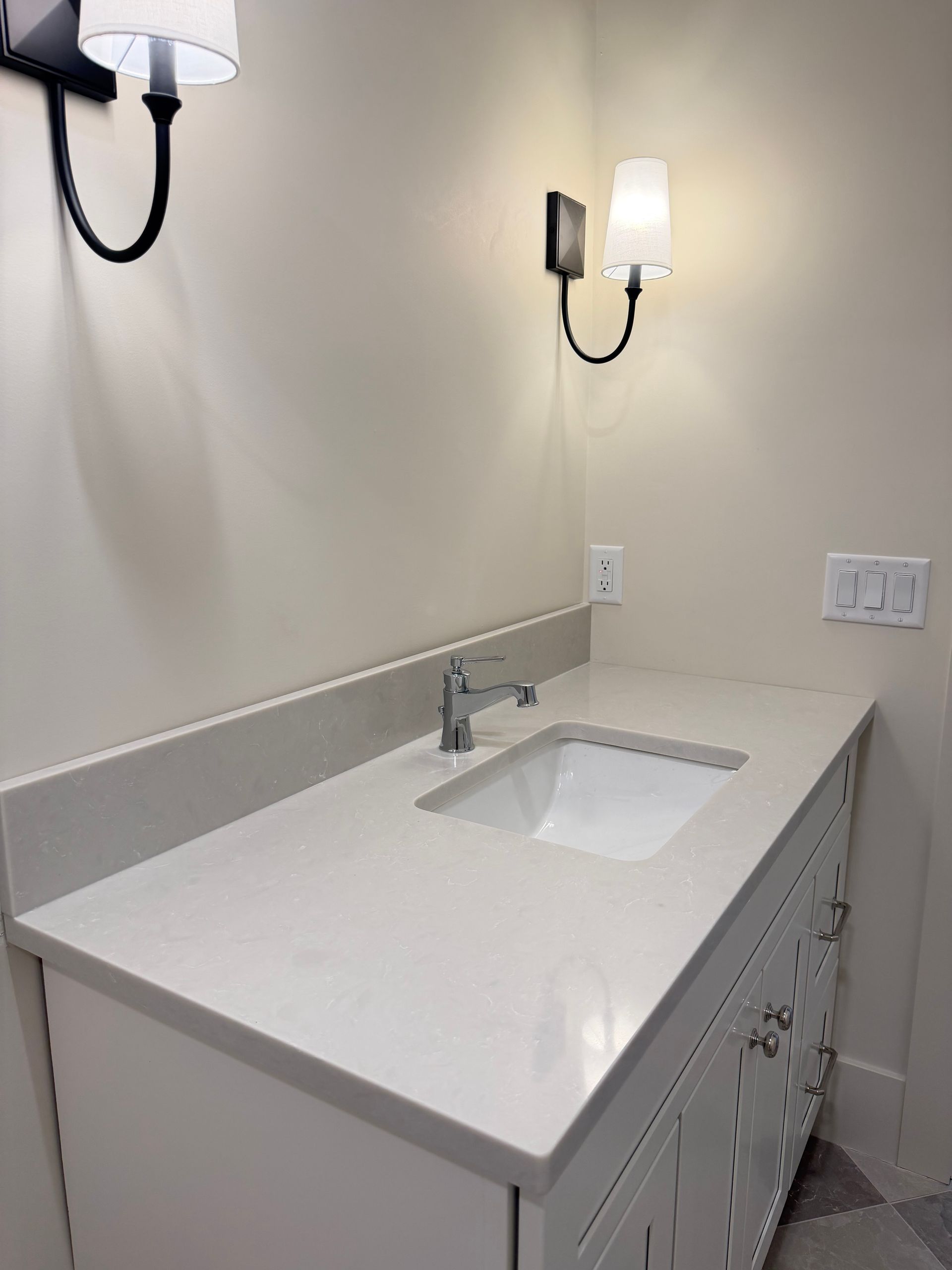 Modern bathroom vanity with white countertop, sink, chrome faucet, and wall sconces above.