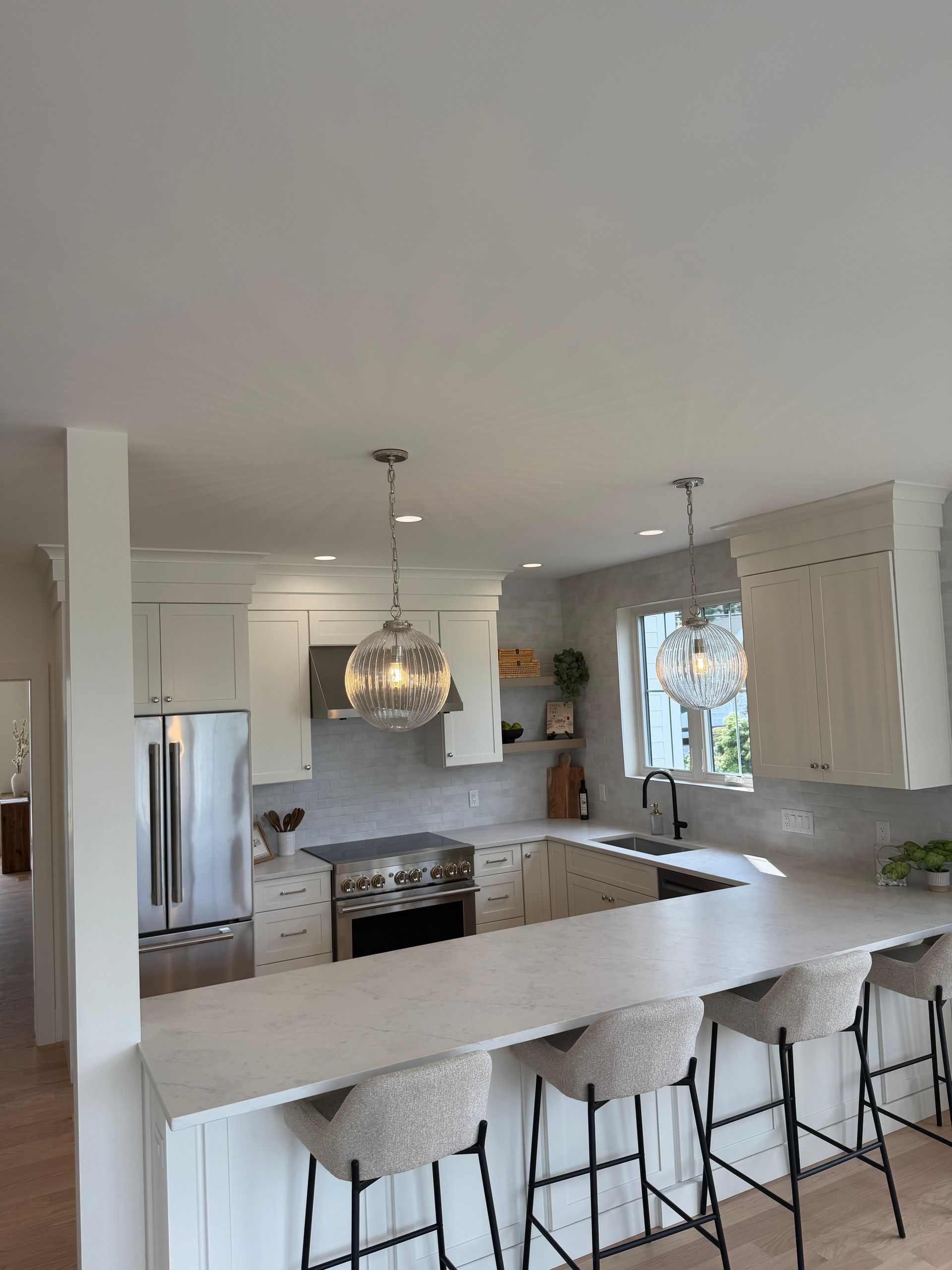 Bright white kitchen with island, pendant lights, stainless fridge, and barstools by a window.