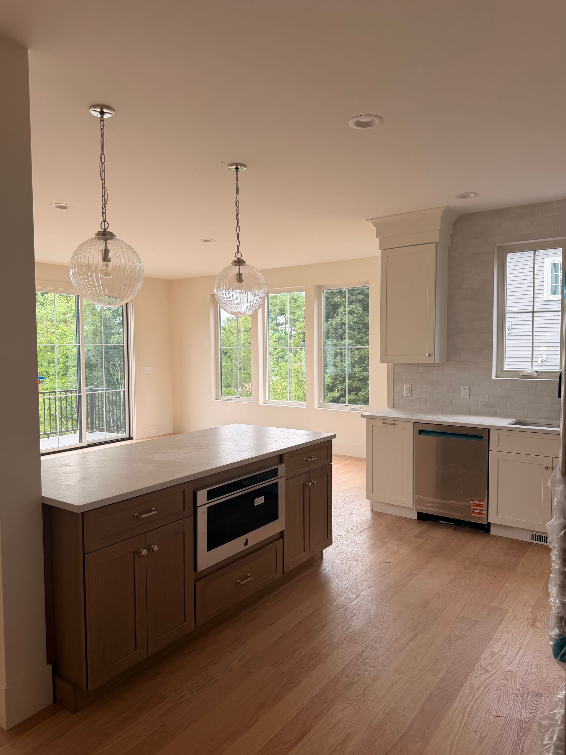 Empty modern kitchen with wood floors, white cabinets, island, pendant lights, and large windows.