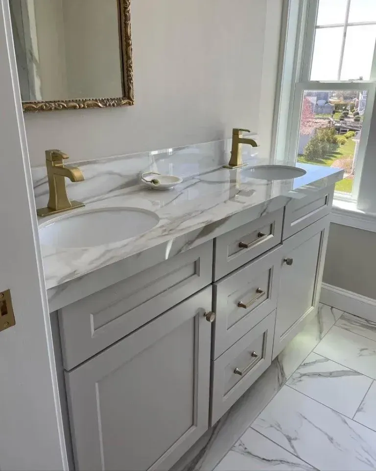 White marble double-sink bathroom vanity with gold faucets beside a sunlit window.