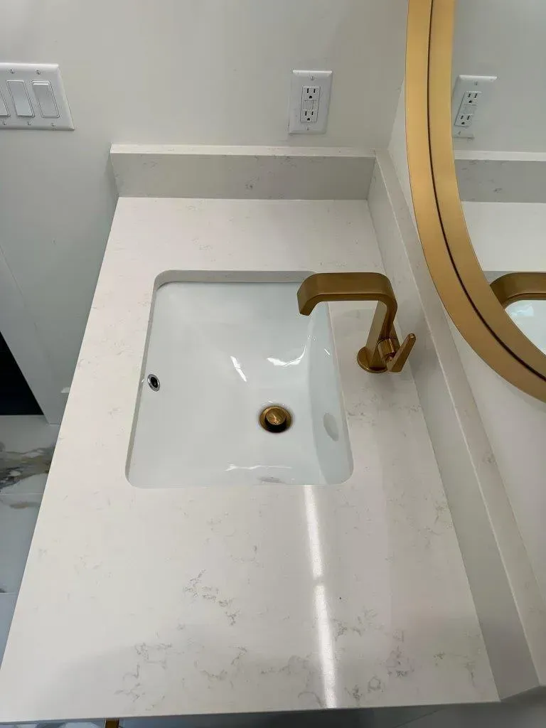 Bathroom sink with white countertop, square basin, and gold faucet near a mirror.
