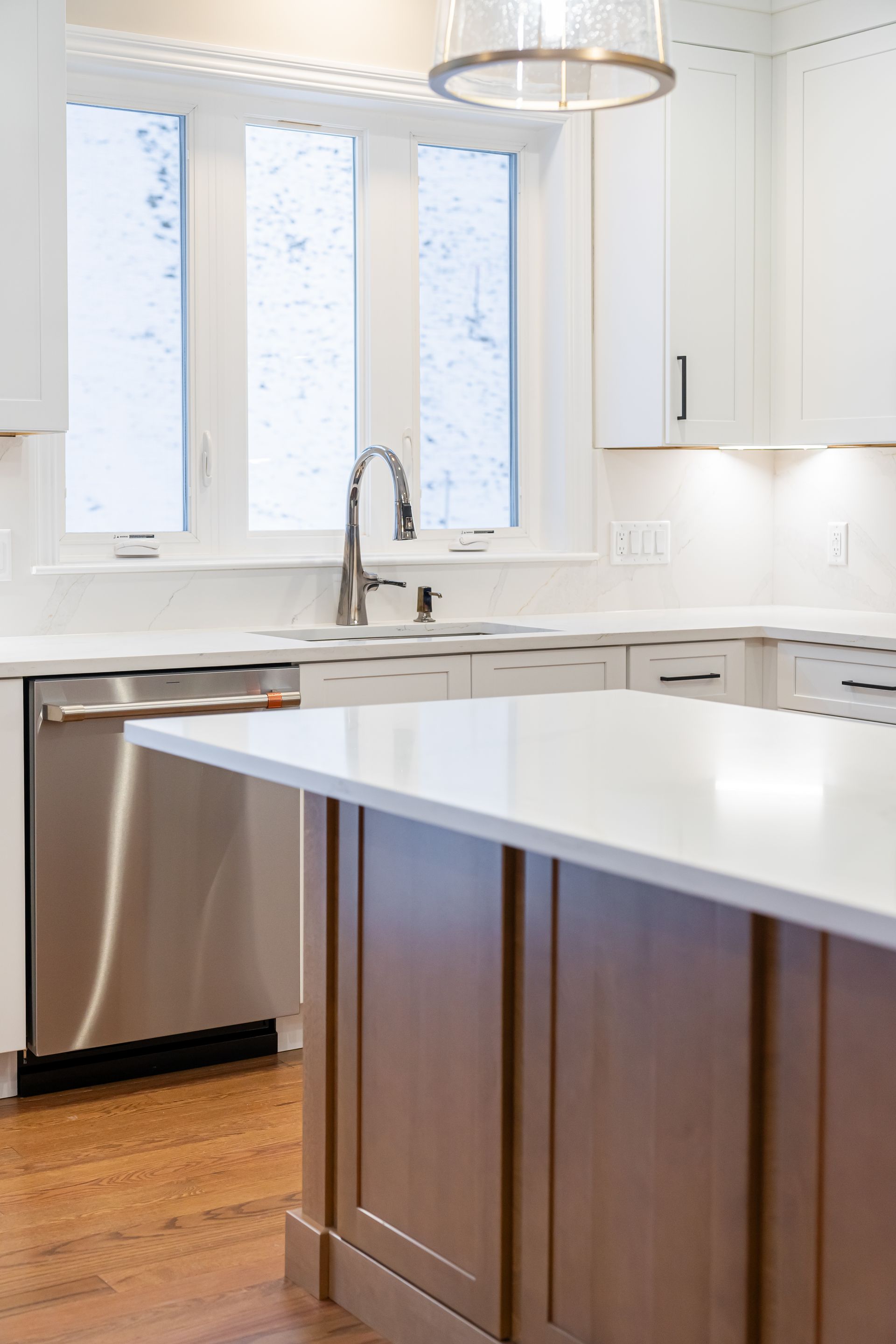 Modern white kitchen with island, stainless dishwasher, wood cabinets, and tall windows.