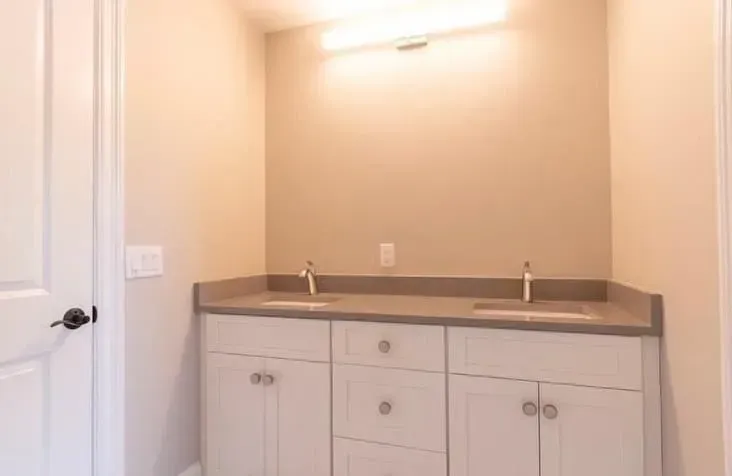 Bathroom vanity with white cabinets, gray countertop, and two sinks beneath a lit mirror