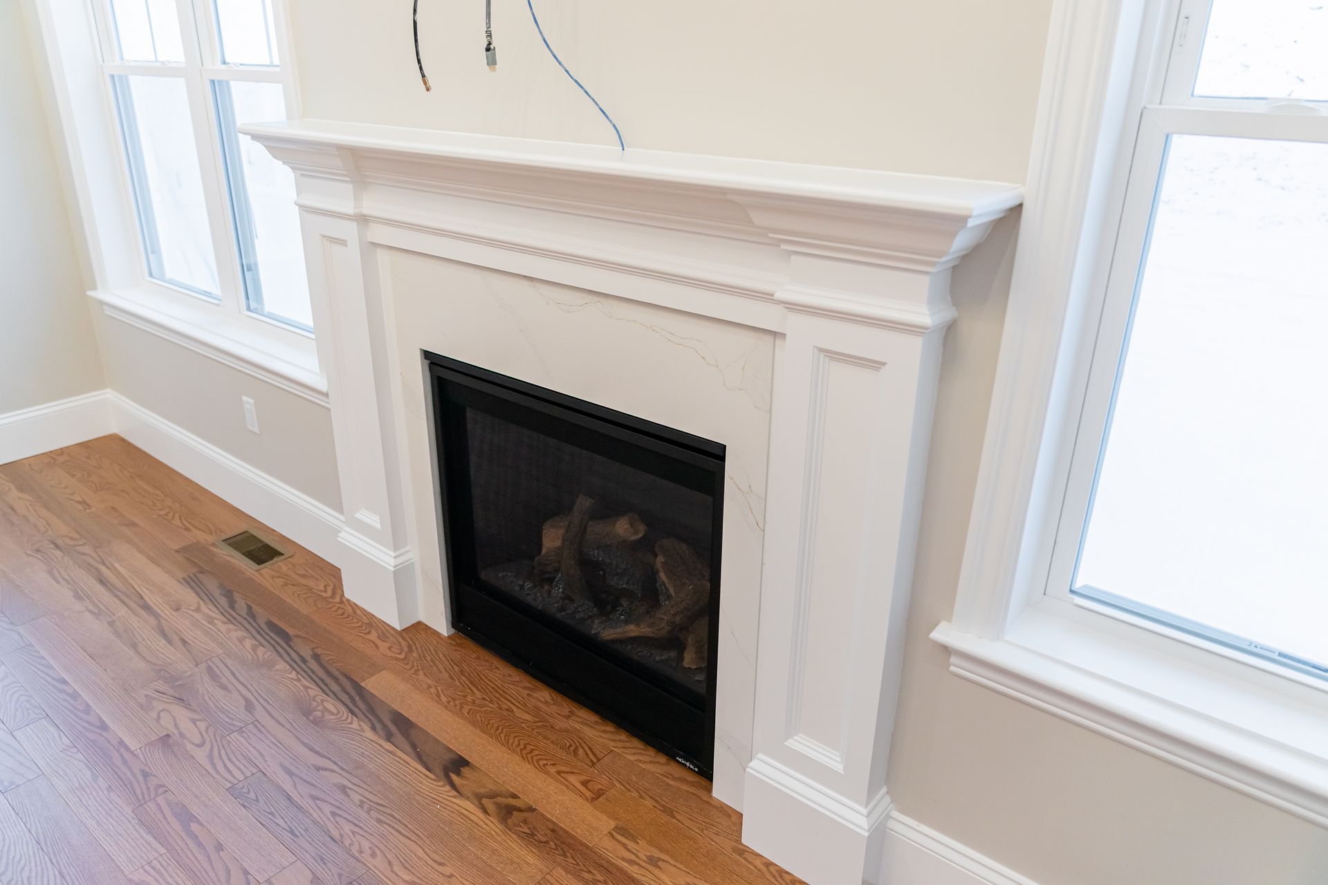 White fireplace with mantel between two windows in a bright room, with hardwood floors and built-in cabinets nearby