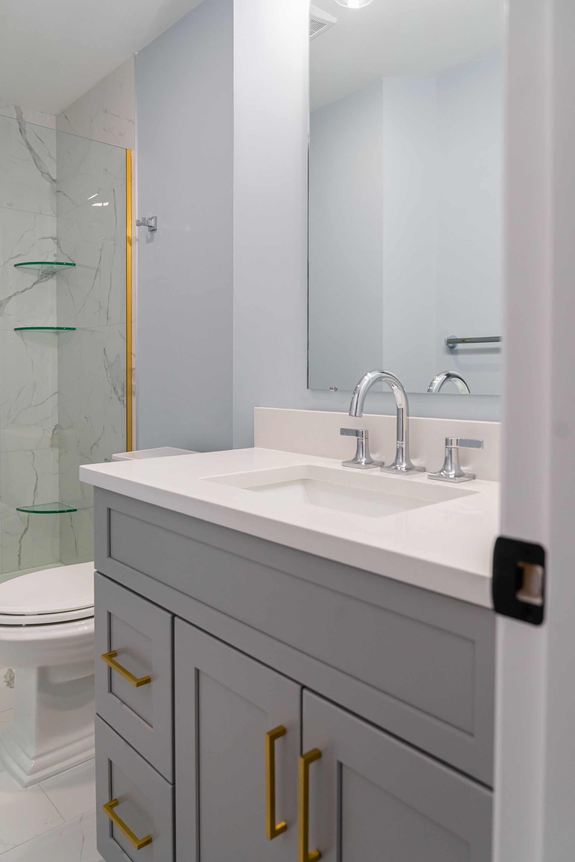 Modern bathroom vanity with gray cabinets, white countertop, and gold hardware beside a glass shower.
