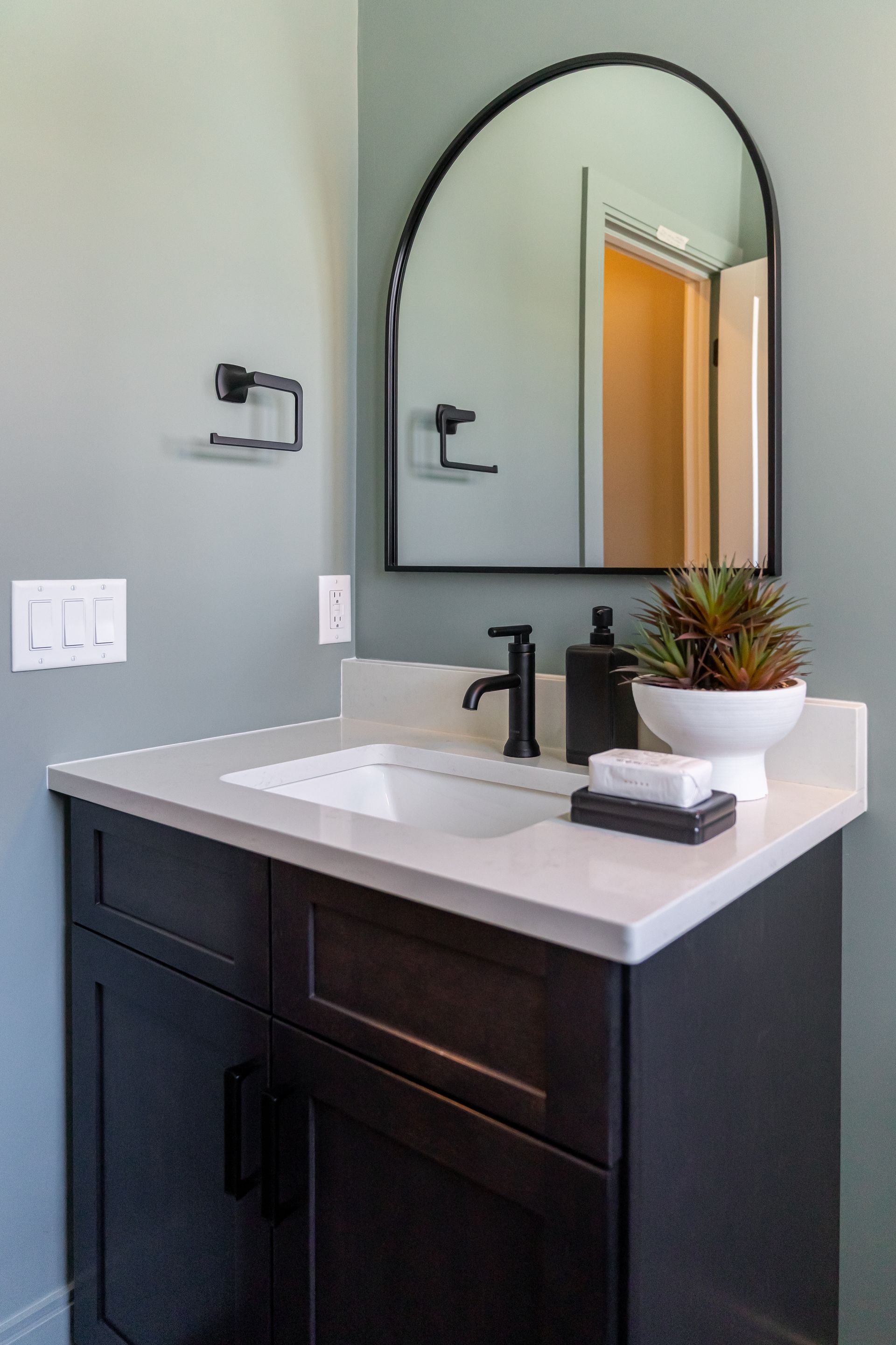 Modern bathroom vanity with black cabinet, white sink, arched mirror, and small plant decor