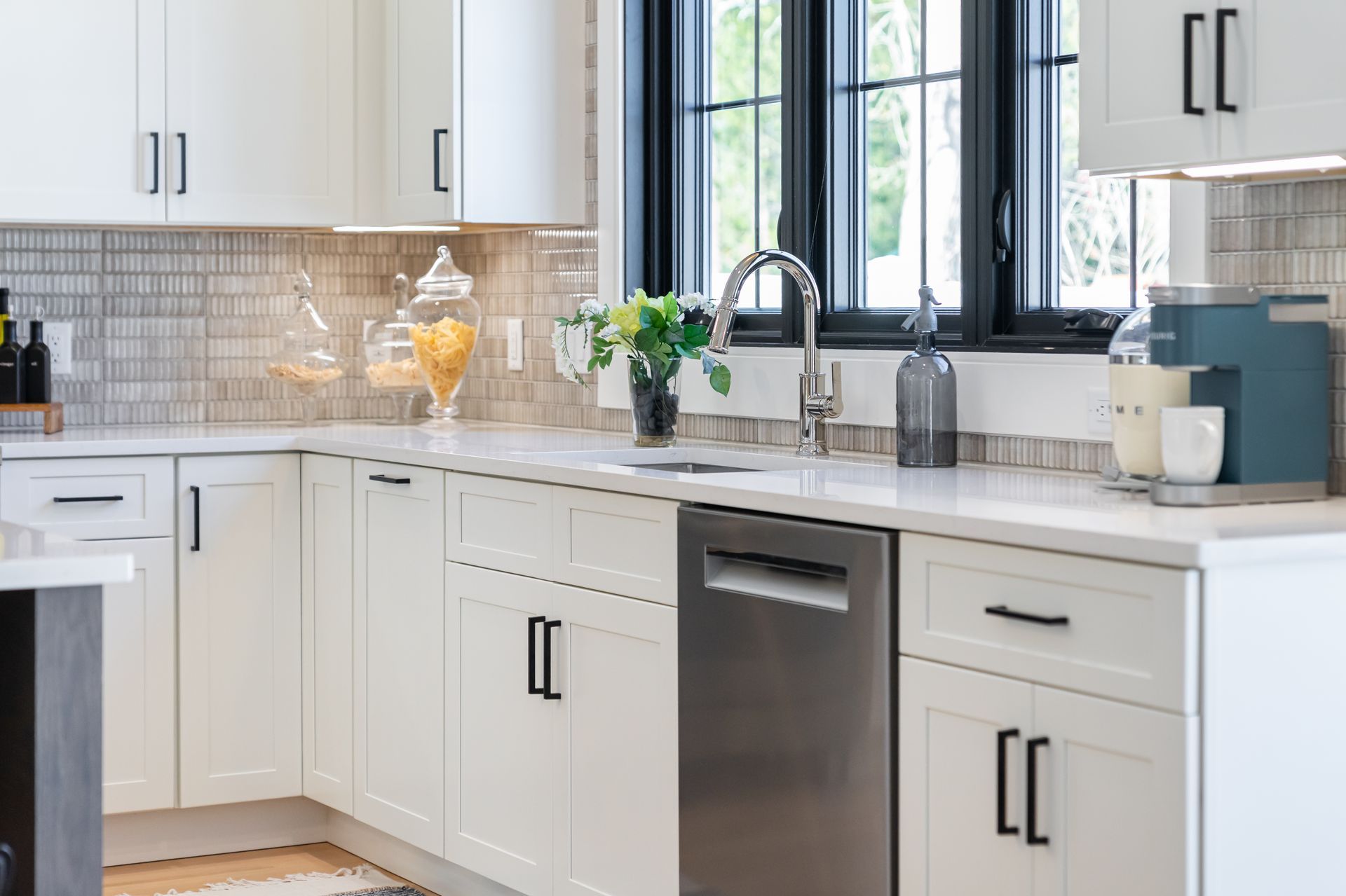 Bright modern kitchen with white cabinets, black hardware, gray tile backsplash, and a large window above the sink