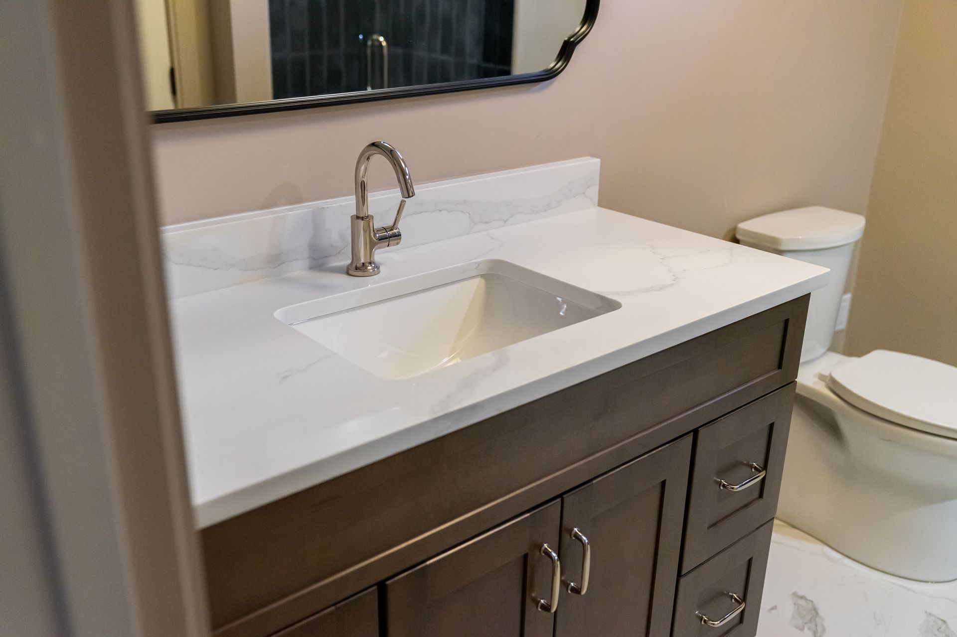 Bathroom vanity with white countertop, dark wood cabinet, sink, faucet, and toilet beside a mirror.