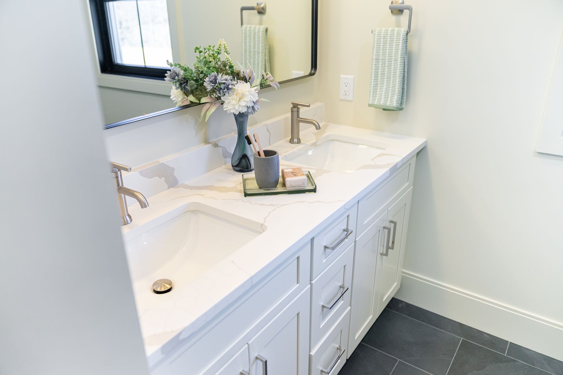 Bright white bathroom vanity with double sinks, mirror, vase of flowers, and toiletries on the counter.