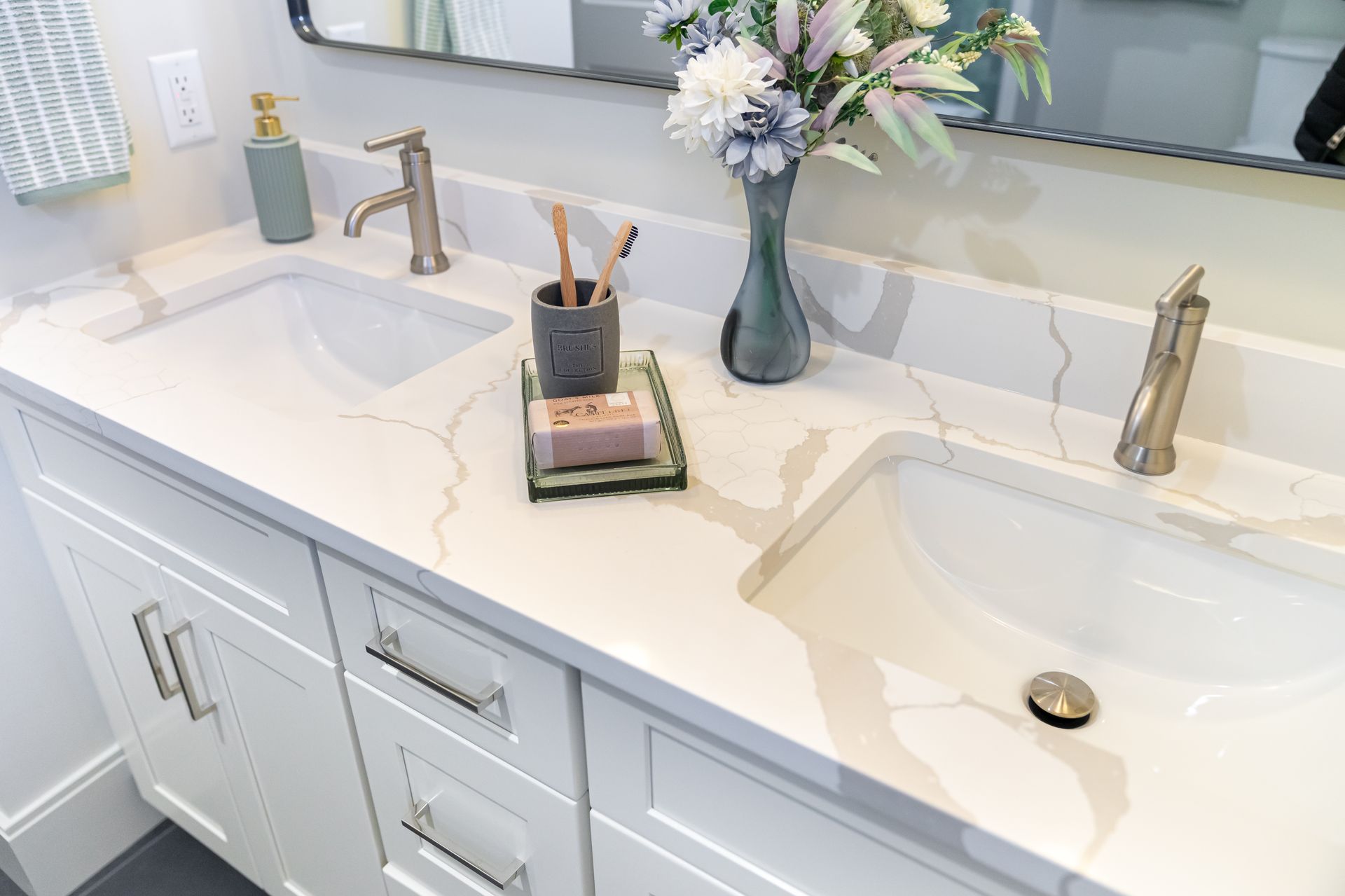 Bright white double bathroom vanity with sinks, gold faucets, marble countertop, and flowers by a mirror