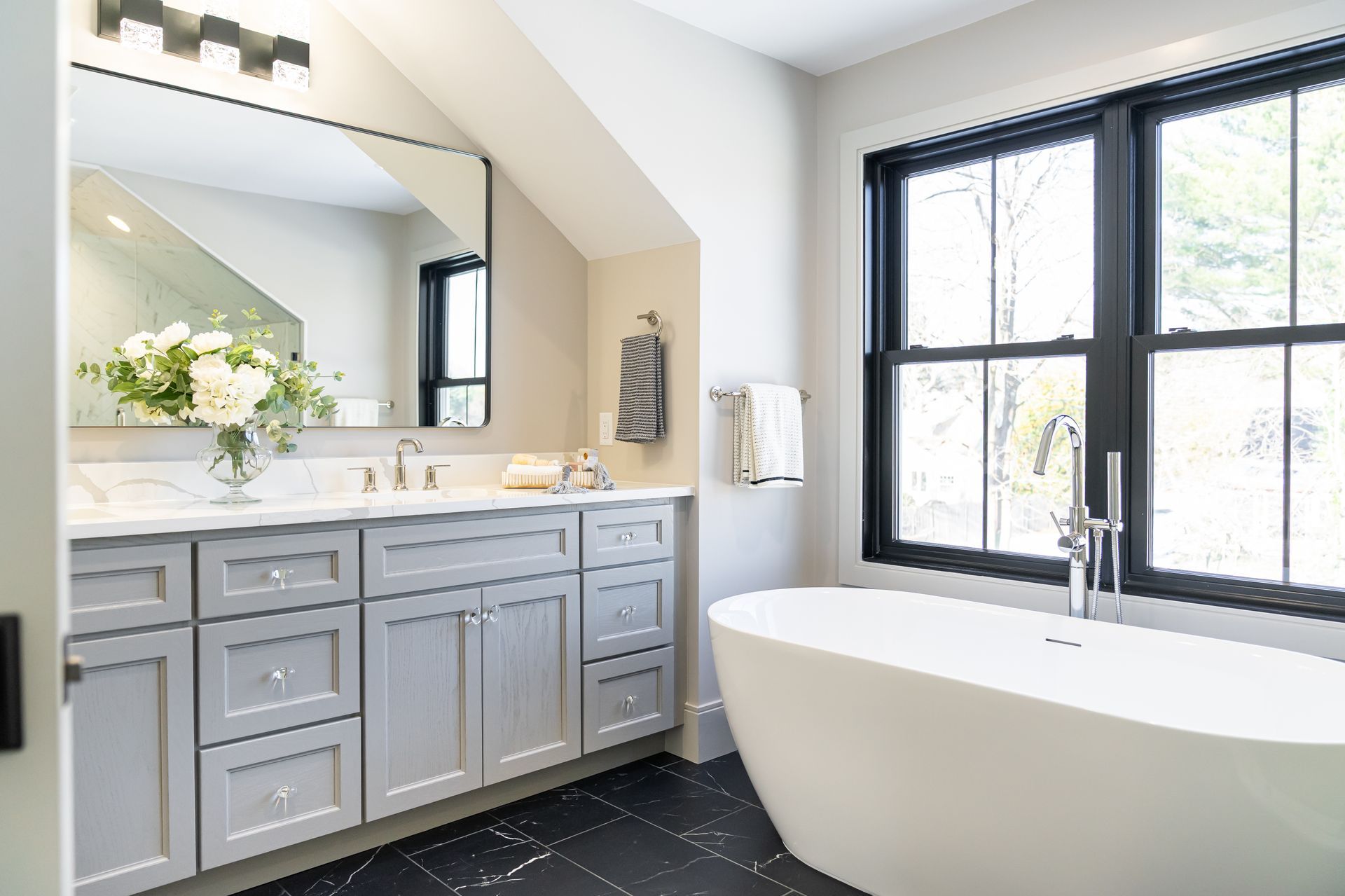 Modern bathroom with gray vanity, large mirror, black-framed windows, and a freestanding white tub
