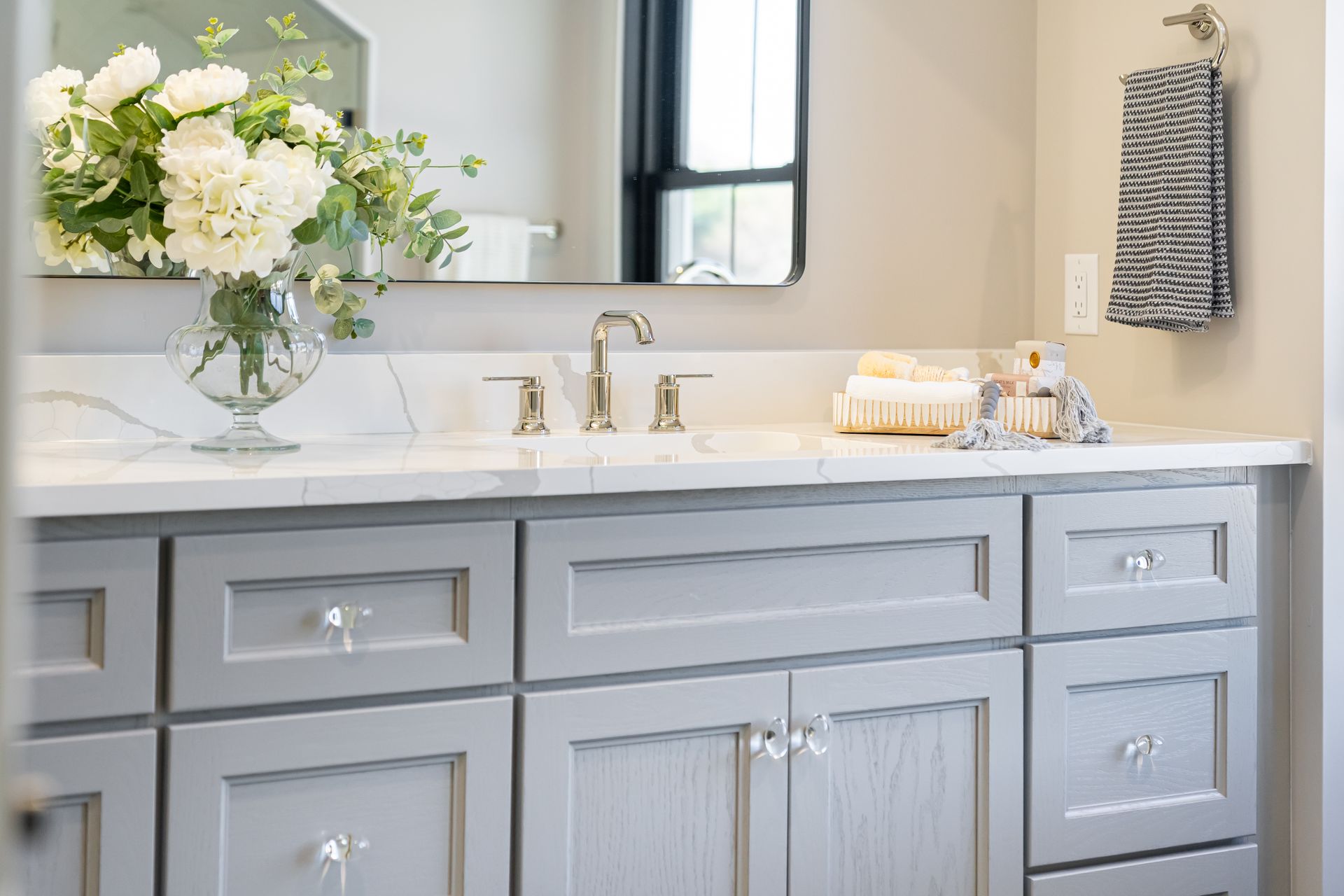 Light gray bathroom vanity with gold fixtures, white flowers, and a striped towel beside a mirror
