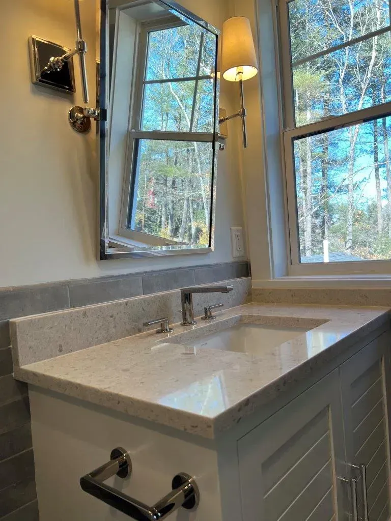 Bathroom vanity with marble countertop, sink, wall sconce, and two windows overlooking trees